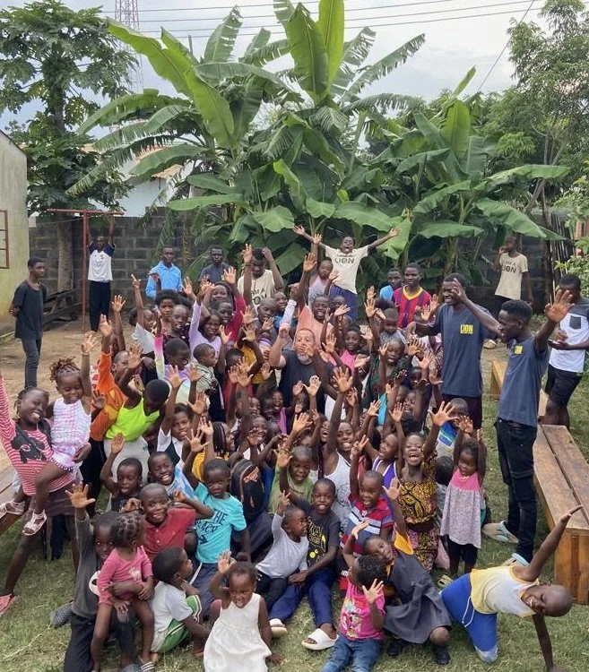 A large group of children and a few adults outdoors, surrounded by lush green banana trees, all smiling and raising their hands in a joyful gathering.