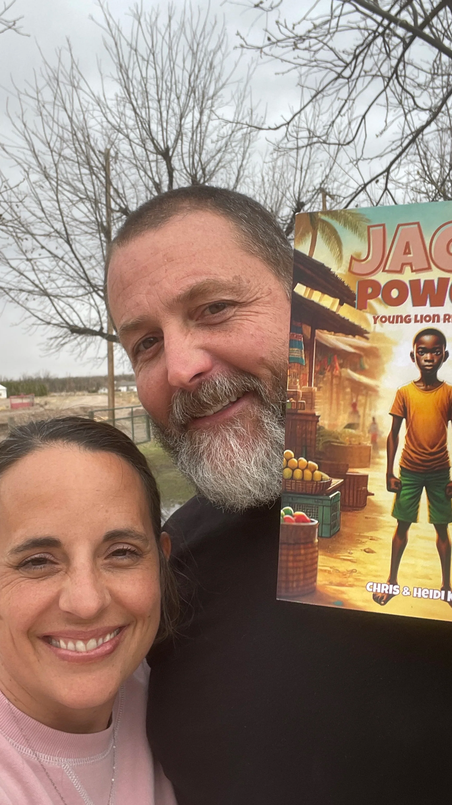 A smiling woman and a smiling man taking a selfie outdoors, with leafless trees and a cloudy sky in the background. The man is holding a book titled 'Jack Power' with a cover illustration of a young boy.