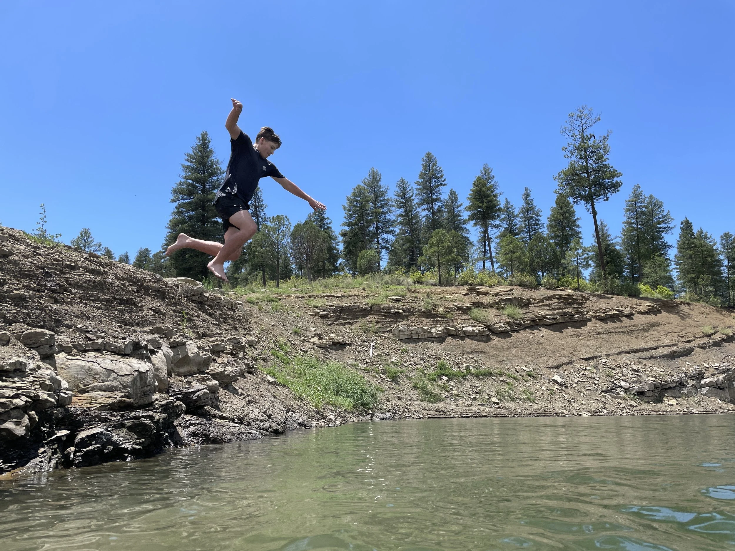 A boy barefoot in black shorts and a t-shirt jumping off a rocky riverbank into a river under a bright blue sky with pine trees in the background.