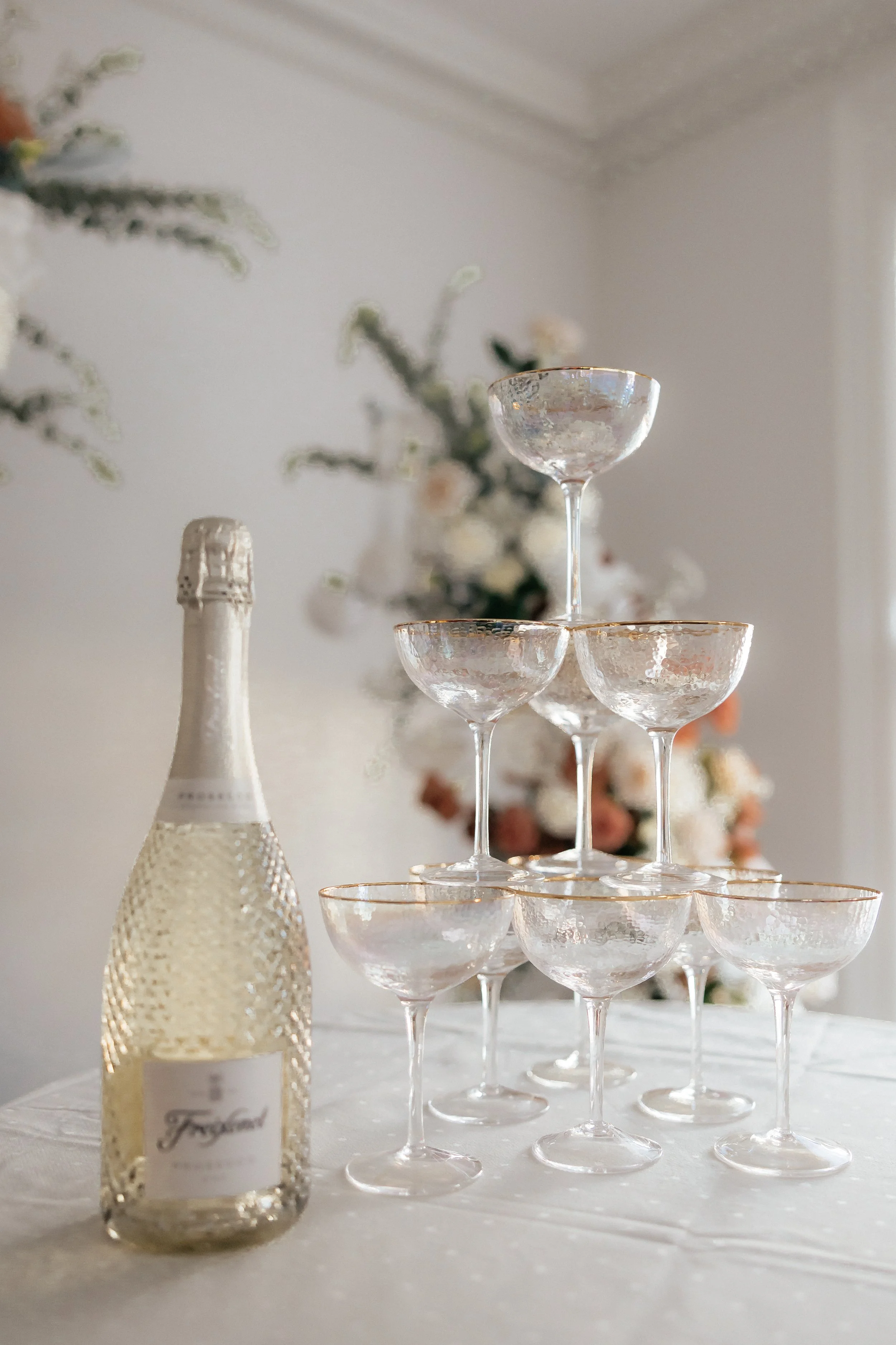 Champagne bottle next to a pyramid of empty coupe glasses on a table with floral decorations in the background.