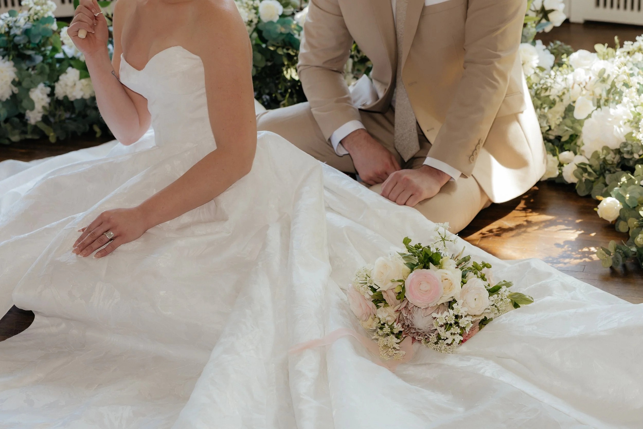 Bride in white gown with bouquet sitting beside a person in a beige suit, surrounded by flowers.
