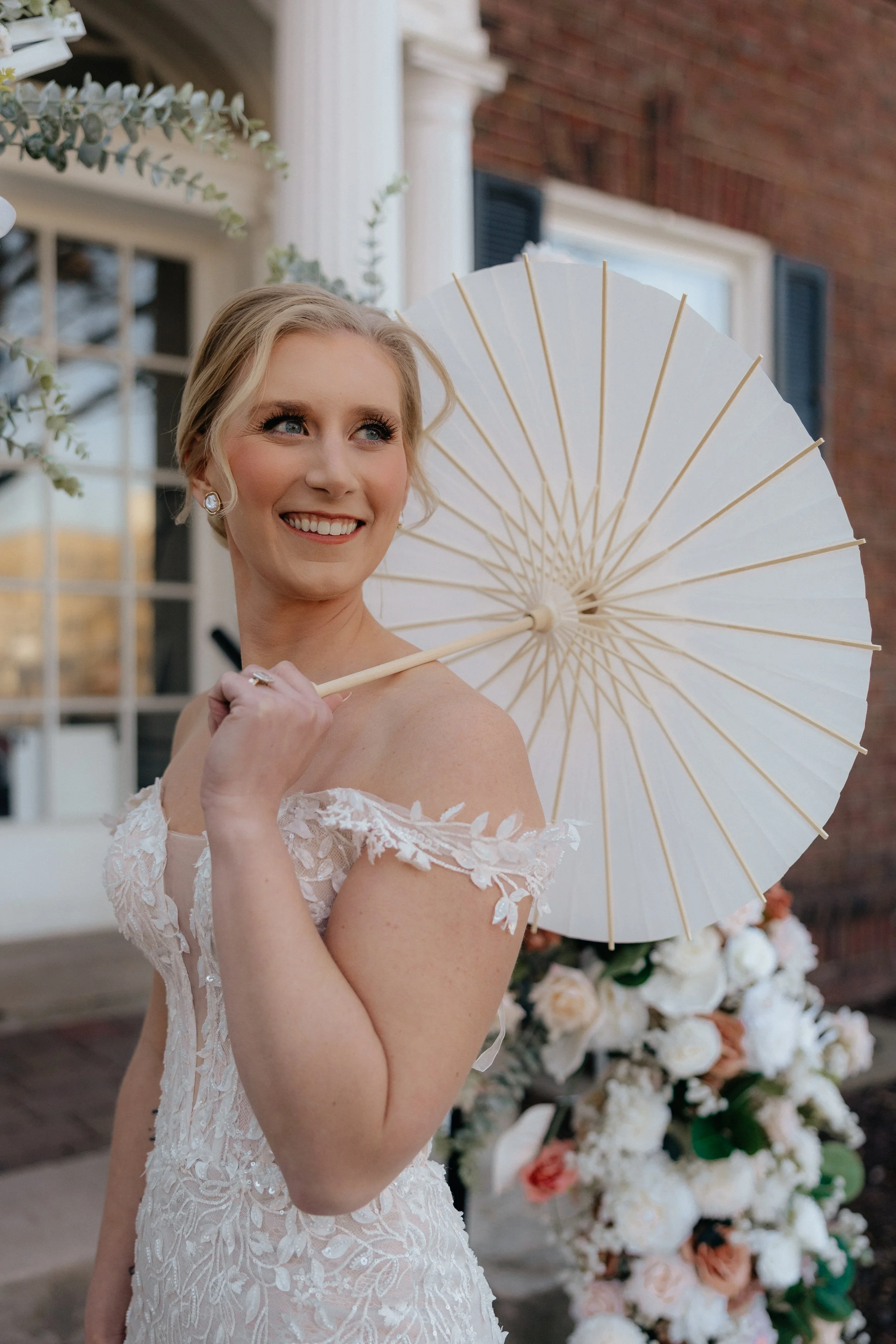 Minneapolis Minnesota Partial Wedding Planning. Woman in a lace wedding dress holding a white paper parasol, smiling, with floral background and brick building.