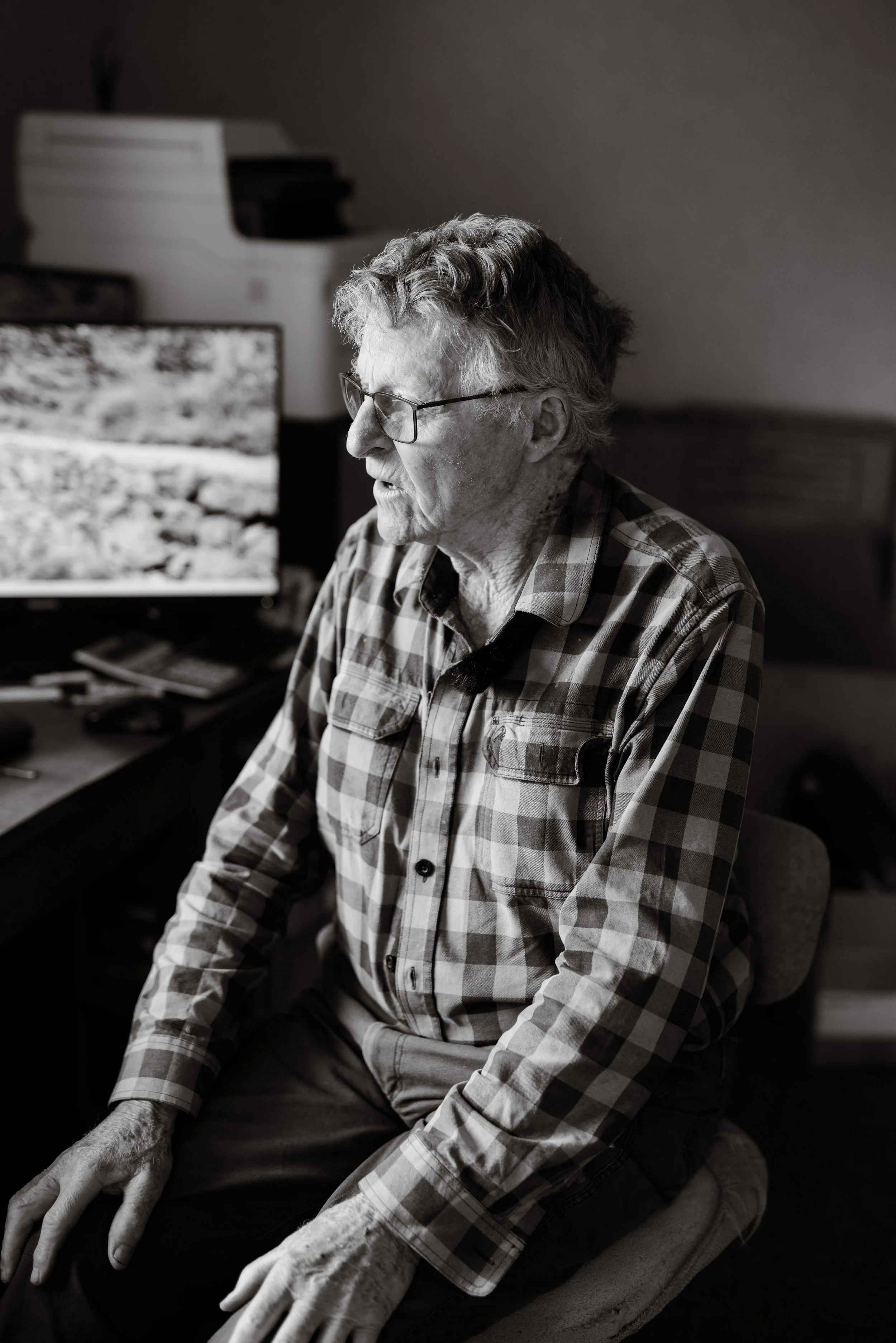 An elderly woman with gray curly hair and glasses is sitting on a chair, wearing a checkered shirt. She appears to be speaking or singing with her eyes closed. In the background, there is a computer monitor displaying a landscape scene and a printer.