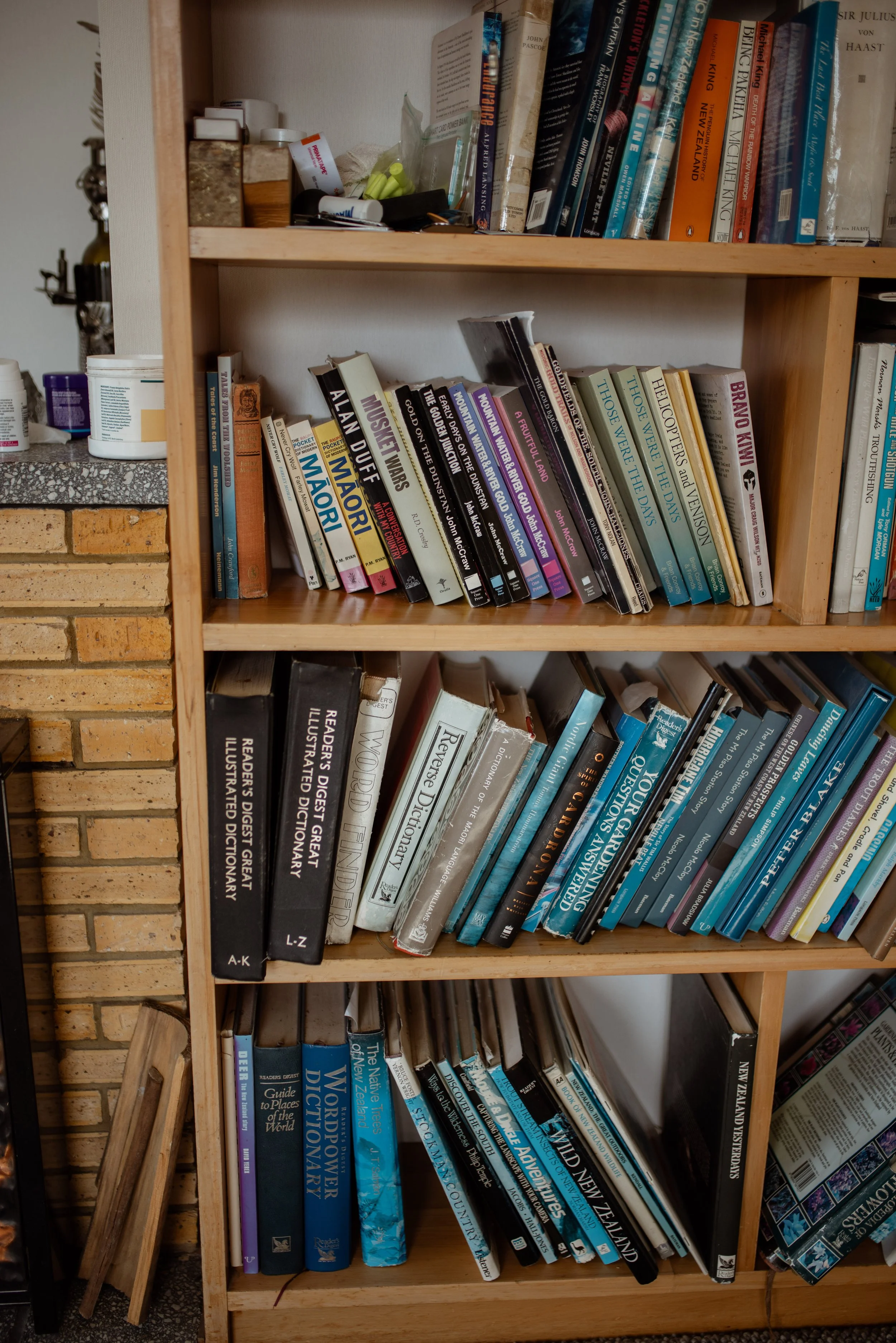 A wooden bookshelf filled with various books, with some books leaning and stacked on top of each other, in a room with brick and drywall walls.