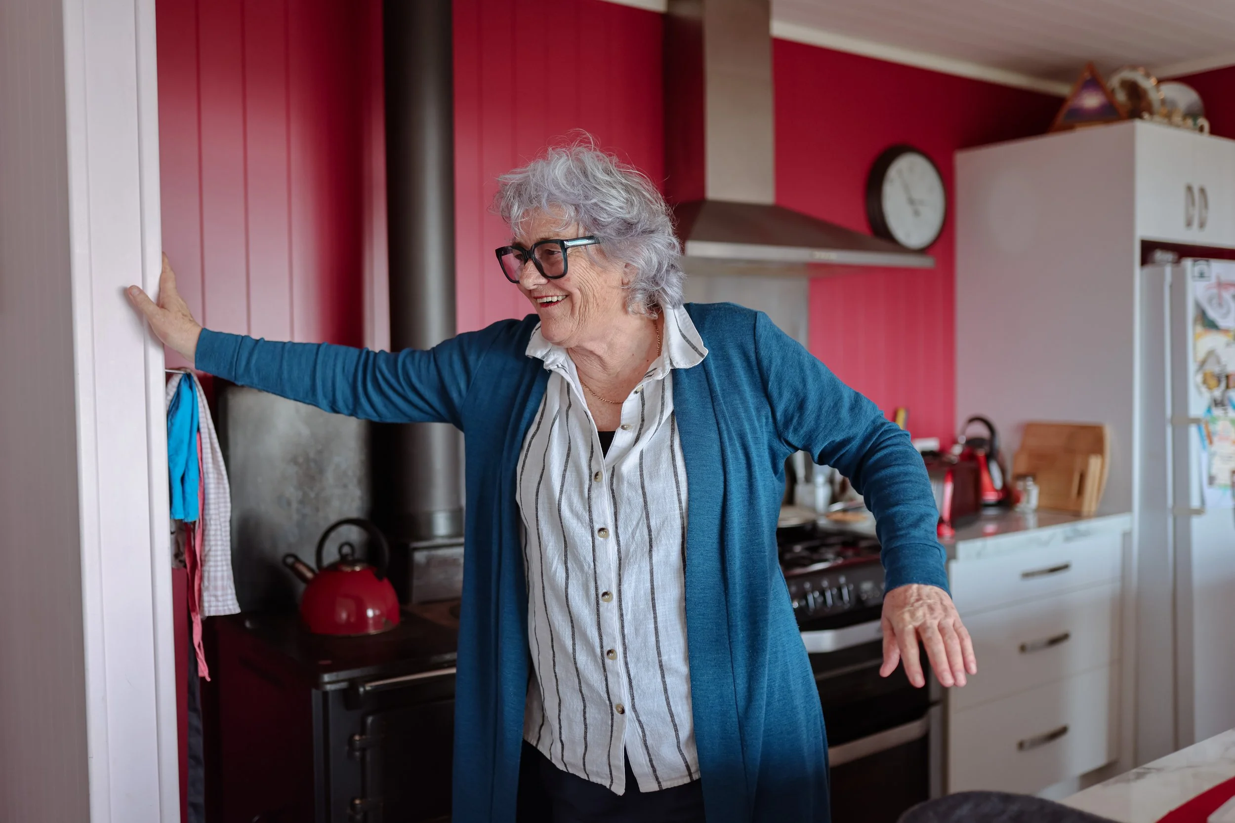 An elderly woman with gray curly hair and glasses smiling cheerfully in a kitchen, extending her arm to the side.