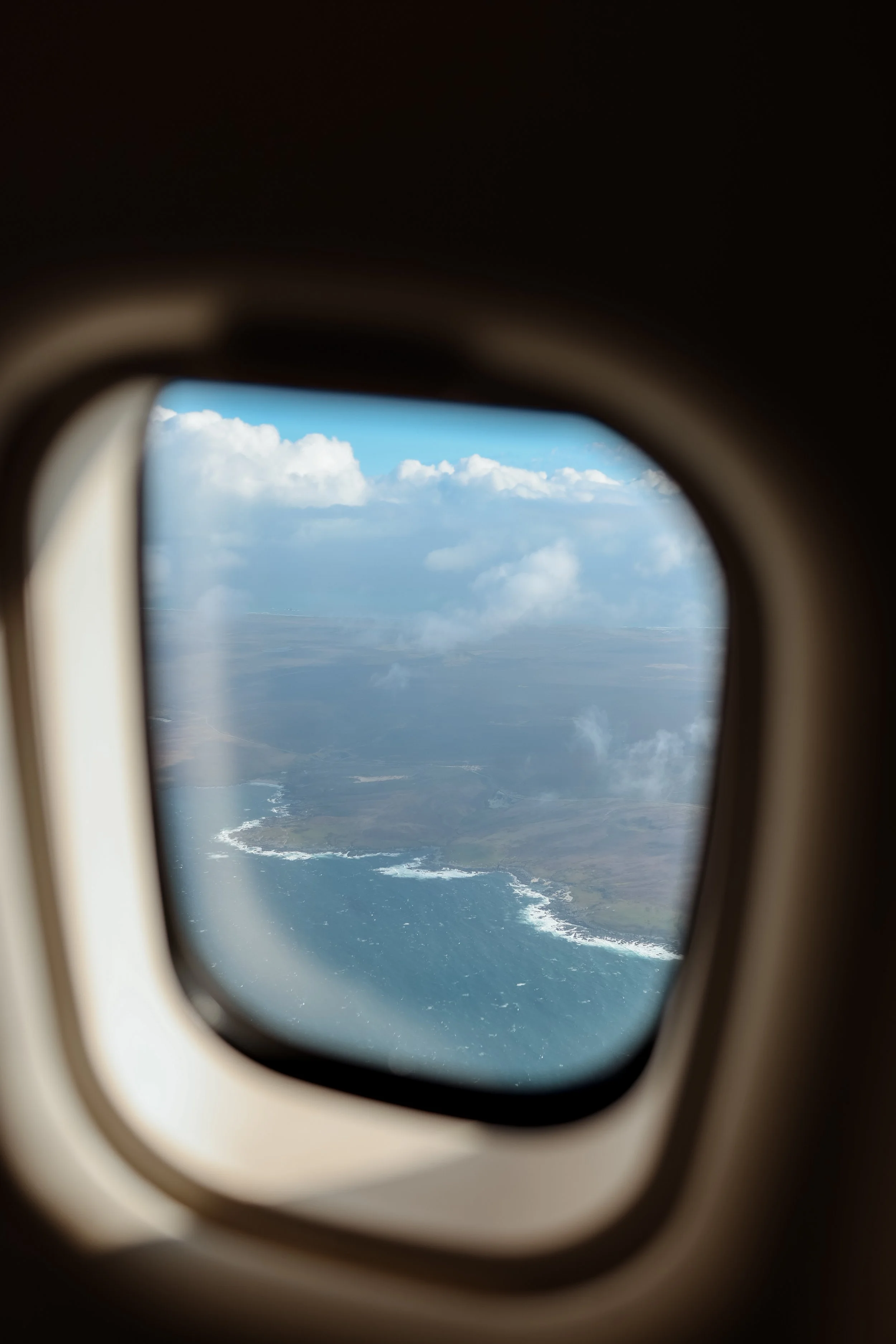 View of a coastline with waves crashing on the shore, seen through an airplane window with clouds in the sky.