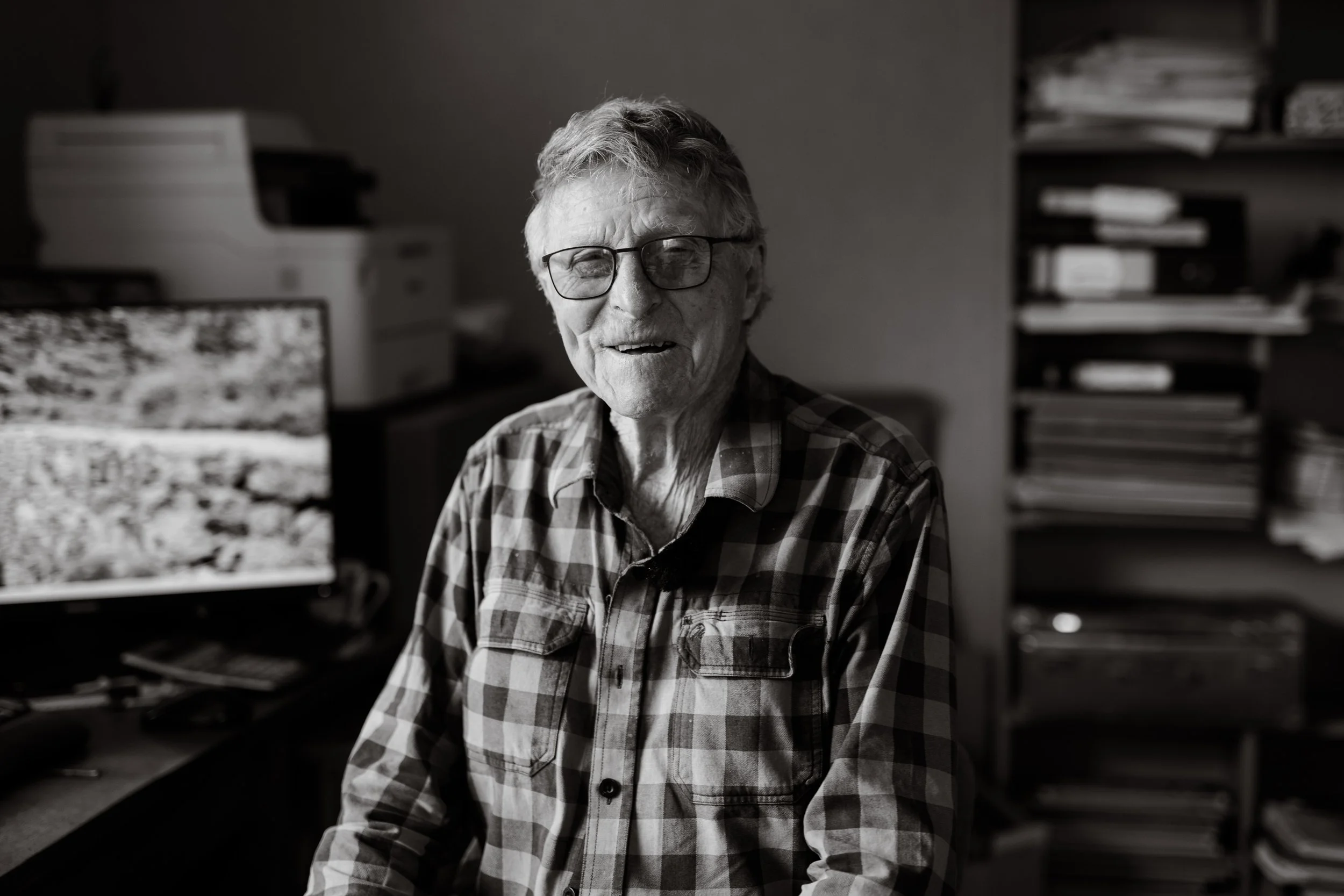 An elderly man with glasses, smiling, wearing a checkered shirt, sitting in an office surrounded by computer equipment and shelves with files.