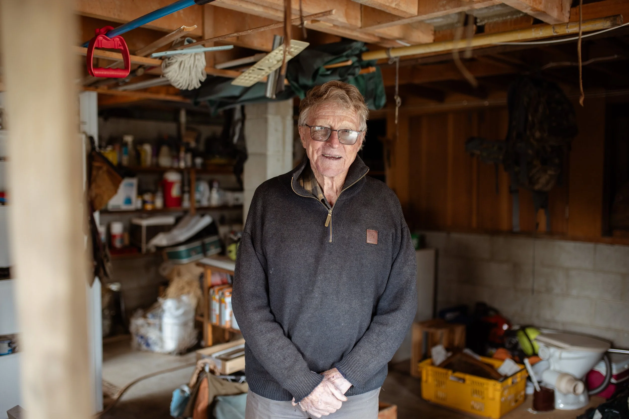 An elderly man with glasses wearing a dark zip-up sweater and gloves stands in a cluttered garage or workshop with shelves filled with various tools, supplies, and objects.