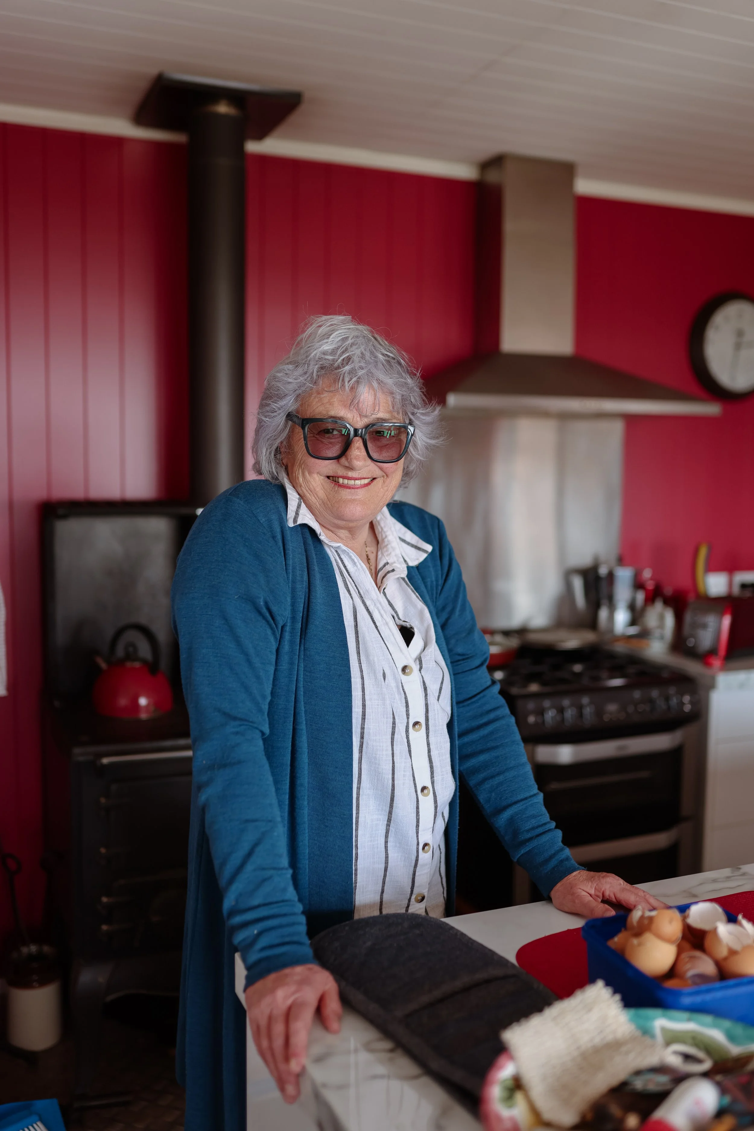 An elderly woman with gray hair, wearing large glasses, a blue cardigan, and a striped shirt, smiling while standing in a kitchen with red walls and a stove in the background.