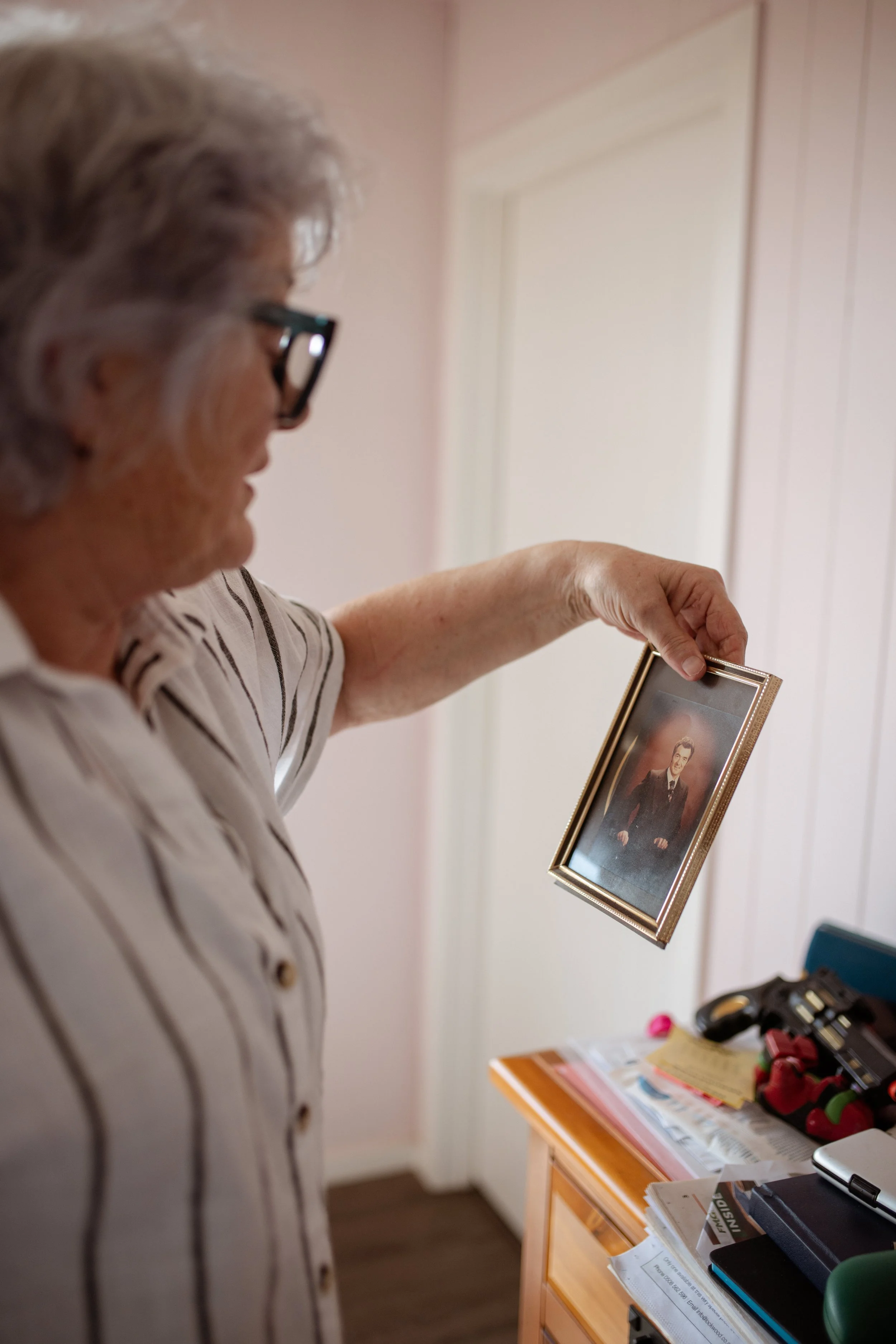 An elderly woman with gray hair and glasses holding a framed photograph.
