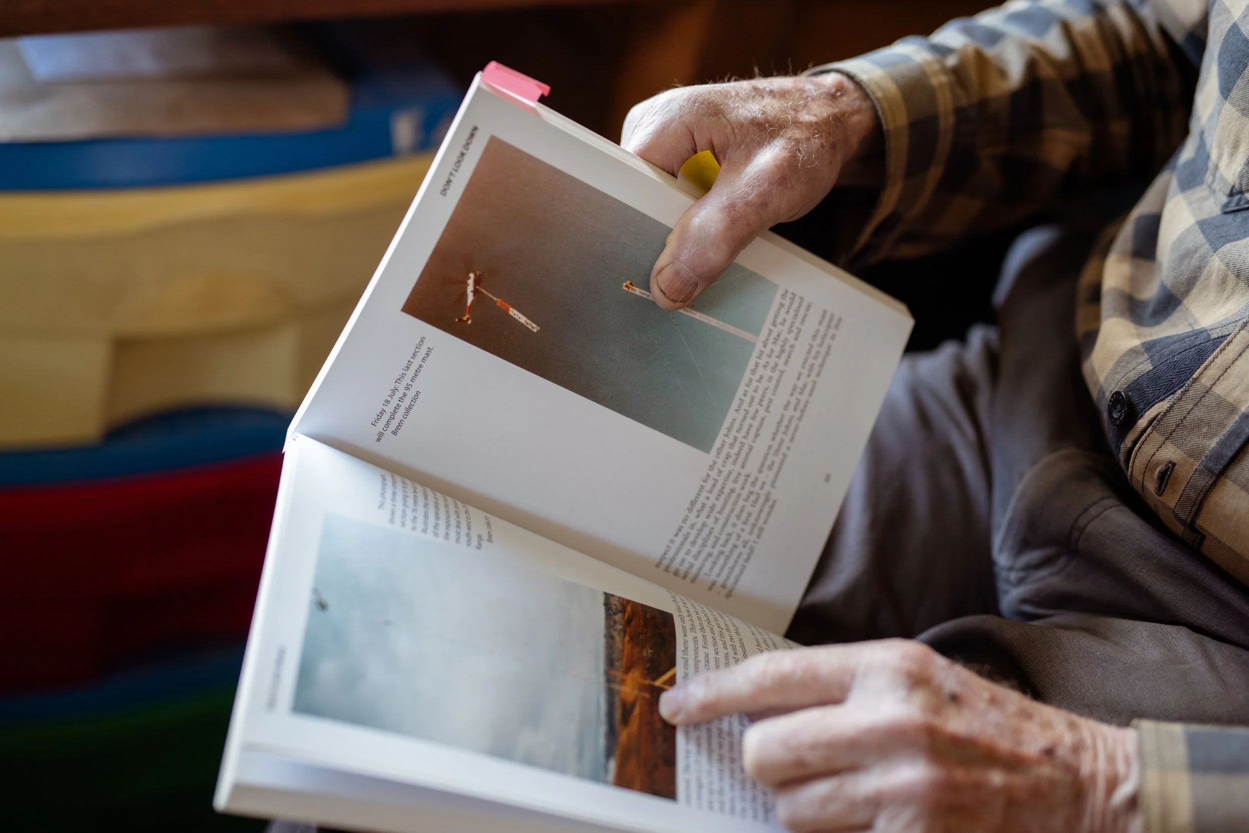 Close-up of an elderly person's hand holding an open magazine, wearing a checkered shirt, sitting at a wooden table.