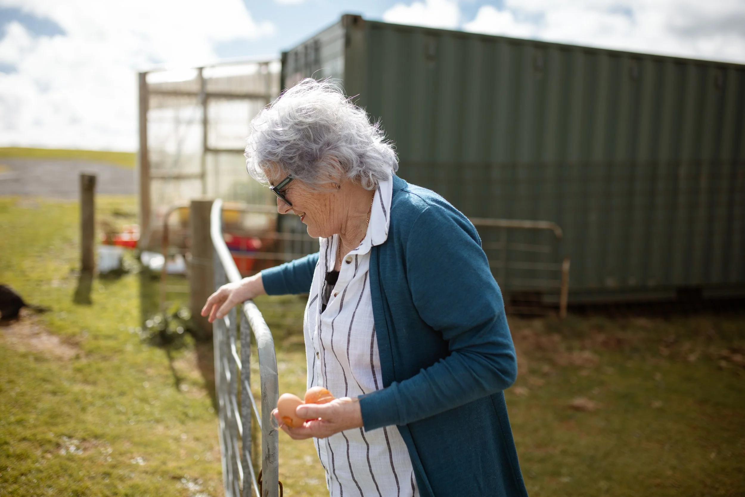 An elderly woman with gray hair and glasses, wearing a white striped blouse and a teal cardigan, stands outside near a fence, holding a couple of eggs.