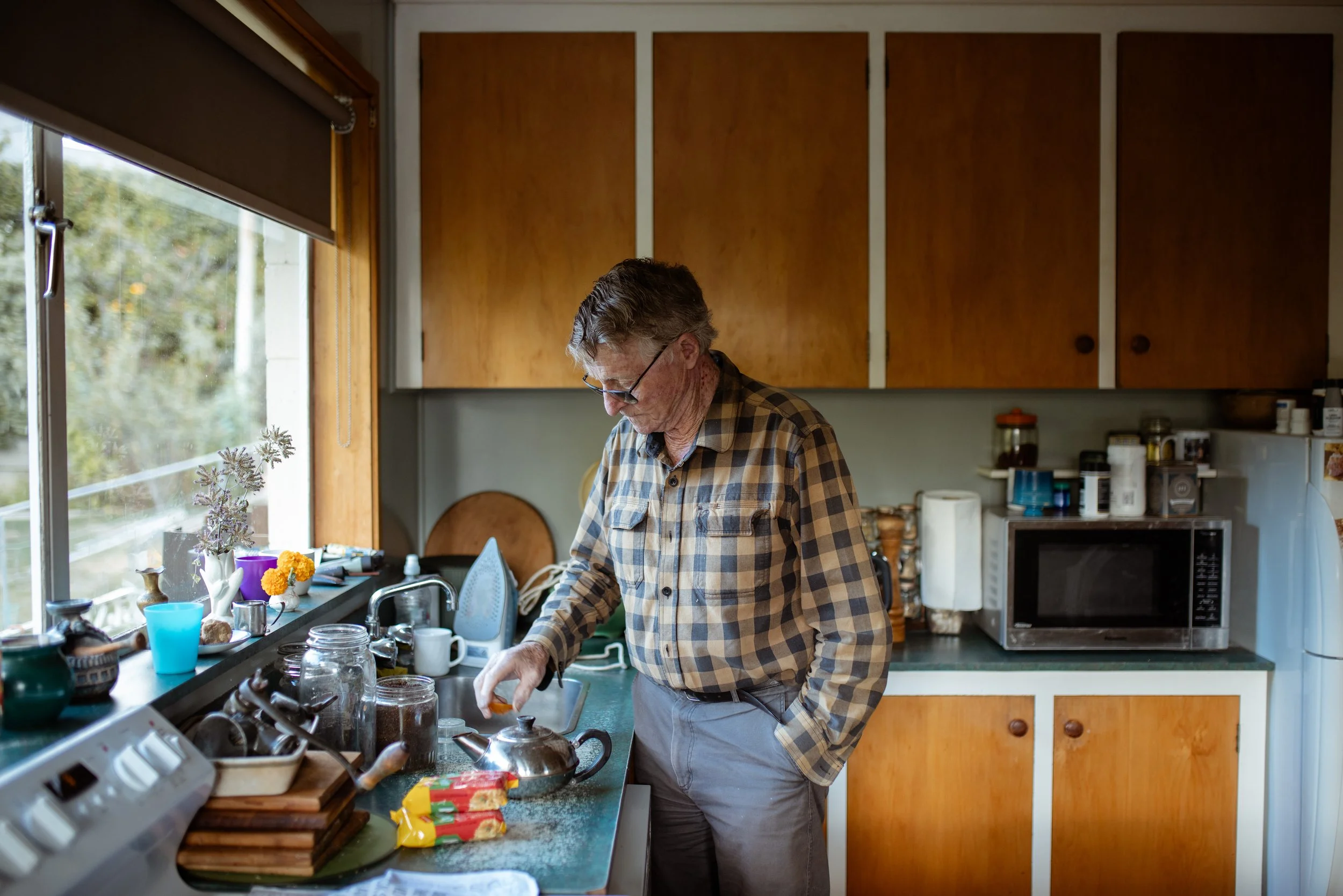 An older man with glasses and a checkered shirt is standing at a kitchen counter, preparing food. The kitchen has wooden cabinets, a window with a view of trees, and various kitchen items and decorations on the counter.