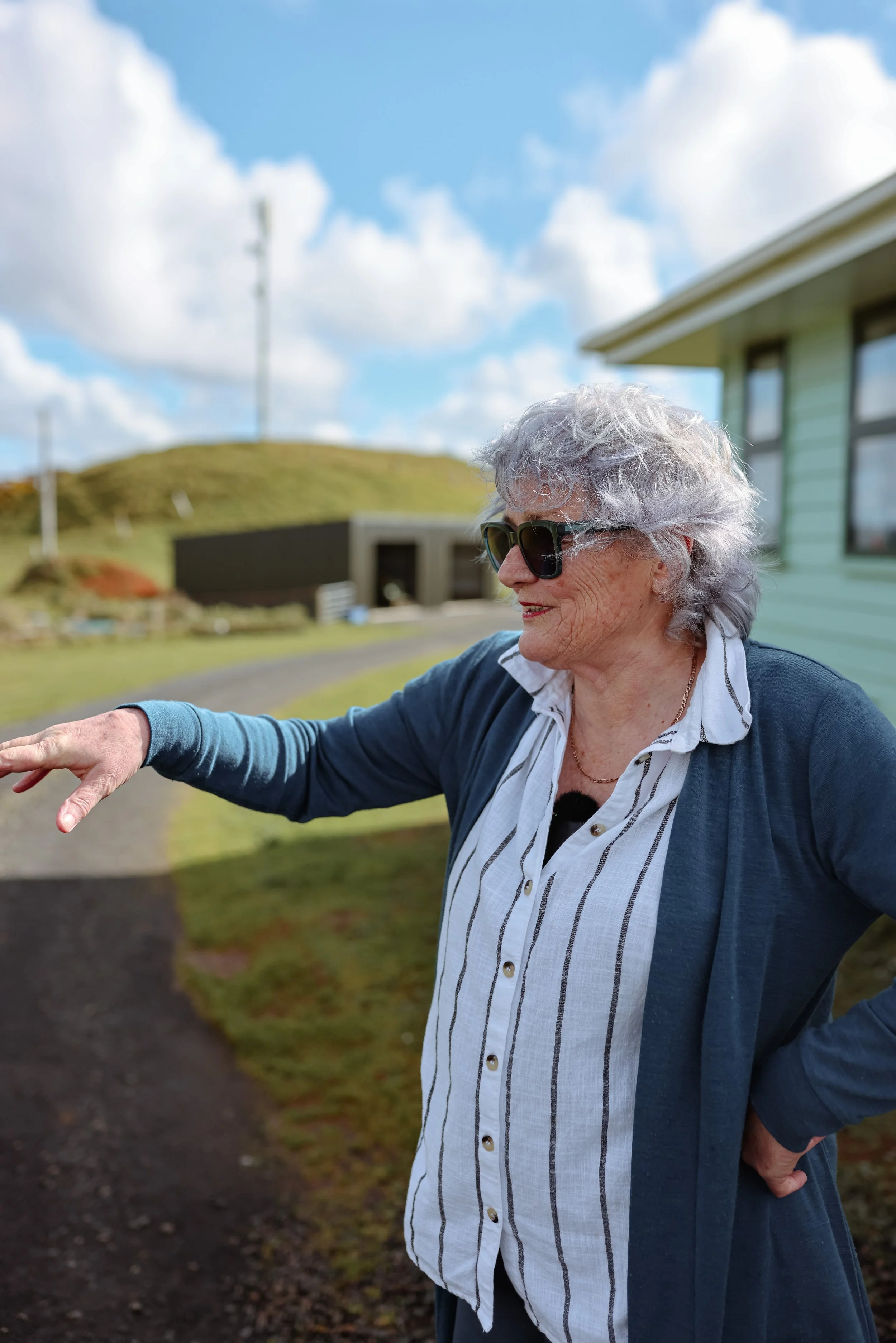 Older woman with gray hair wearing sunglasses, a white button-up shirt, and a dark blue cardigan, standing outdoors on a sunny day with a house and grassy hill in the background.