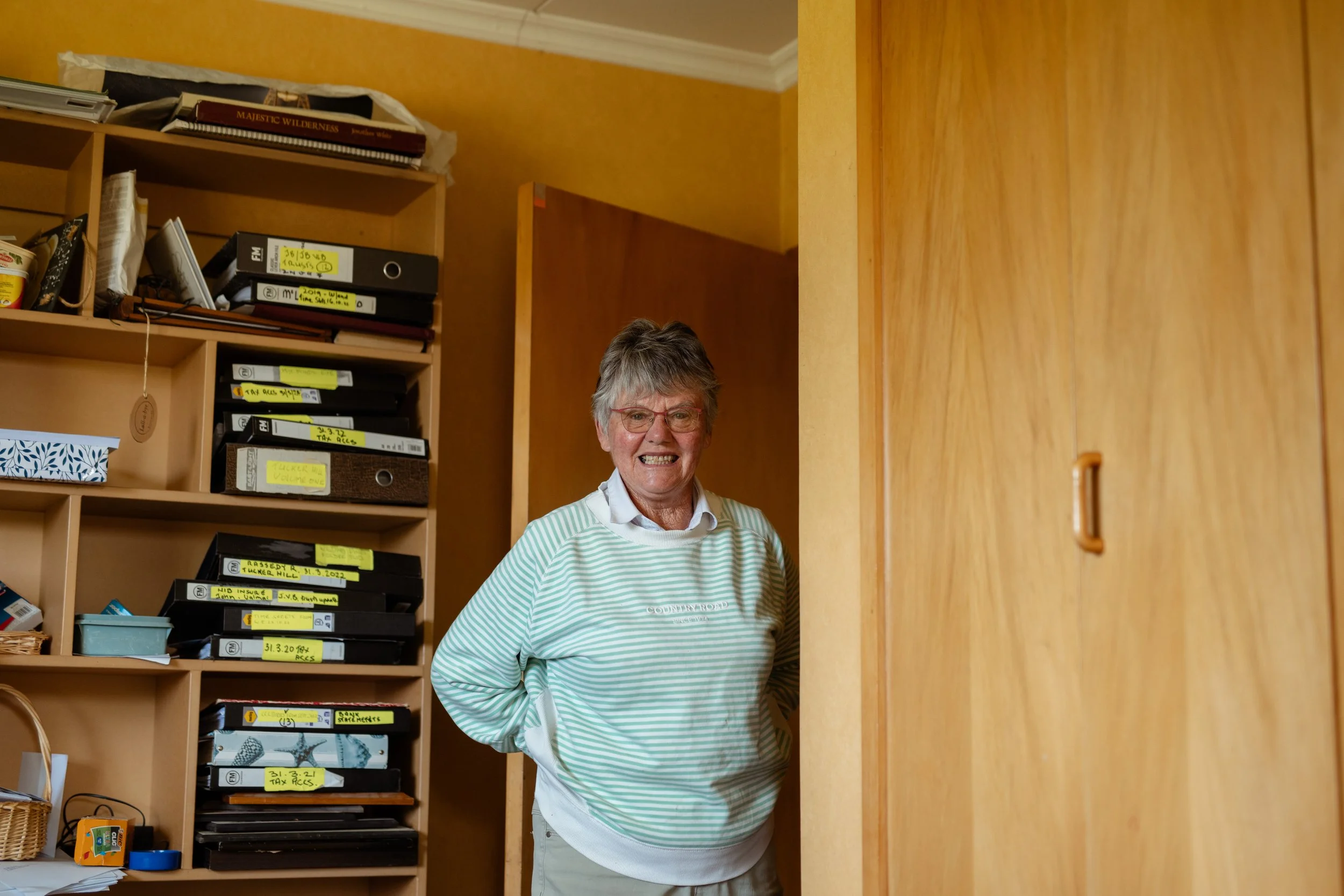A smiling elderly woman with gray hair and glasses standing in a room with wooden shelves filled with binders, books, and miscellaneous items, next to a light wooden door.