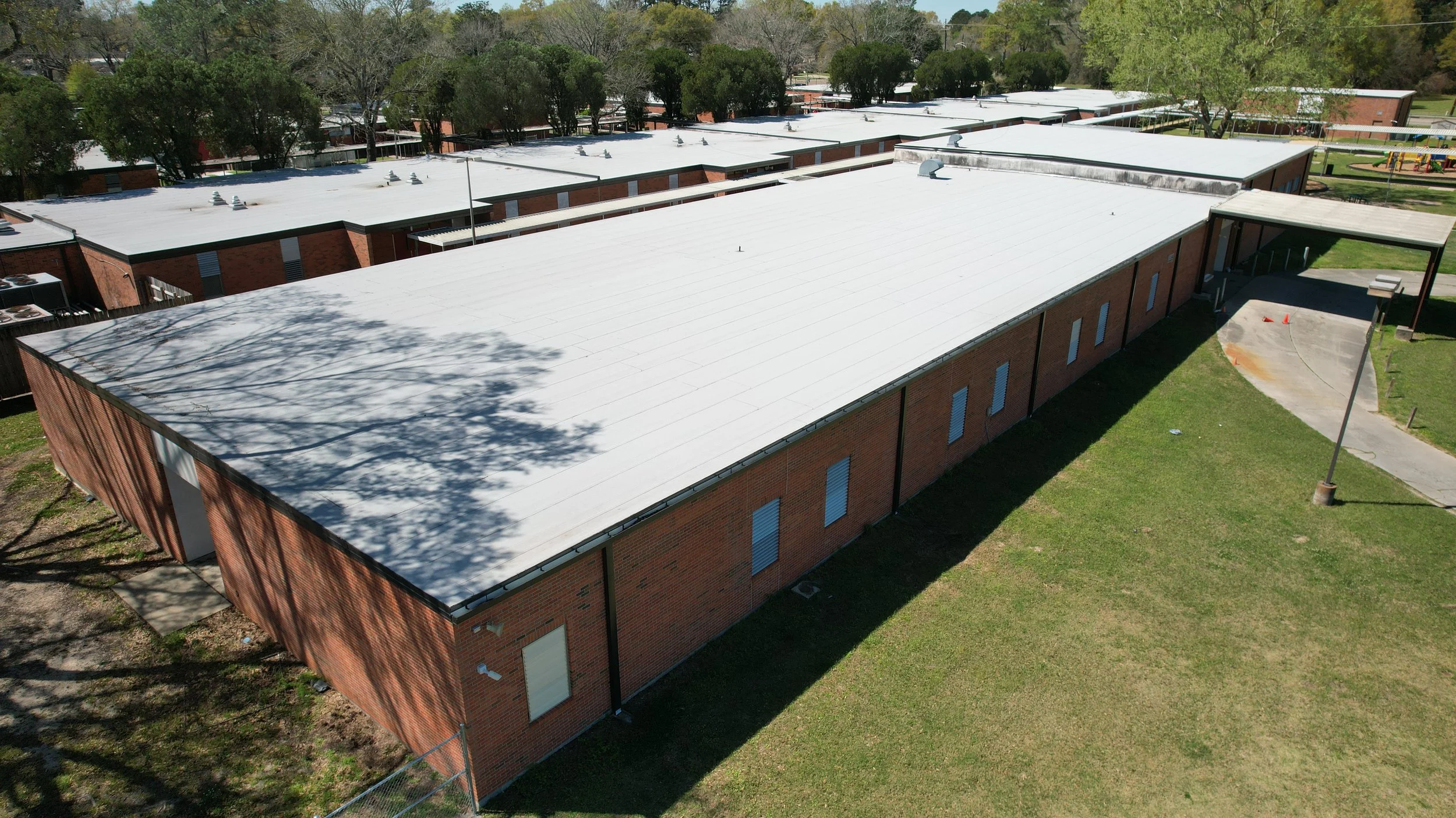 Aerial view of a school building with a newly installed white roof, brick walls, and surrounding grassy areas, trees in the background, and a small playground visible in the distance.