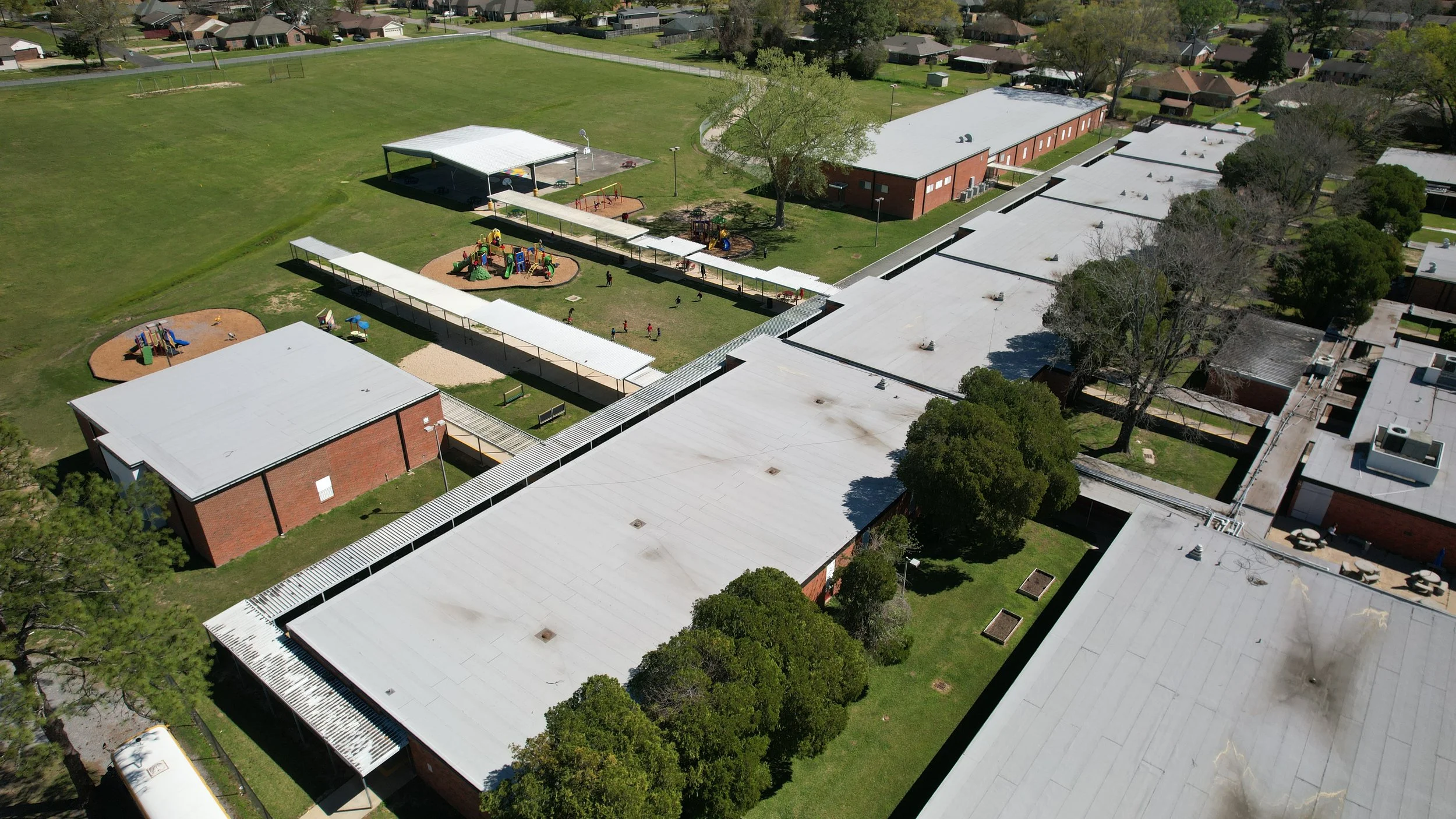 Aerial view of a school campus with multiple school buildings, playgrounds, and green fields, surrounded by trees and residential houses. Each building has a newly installed white panel roof constructed by Supreme Roofing and Construction from Kenner
