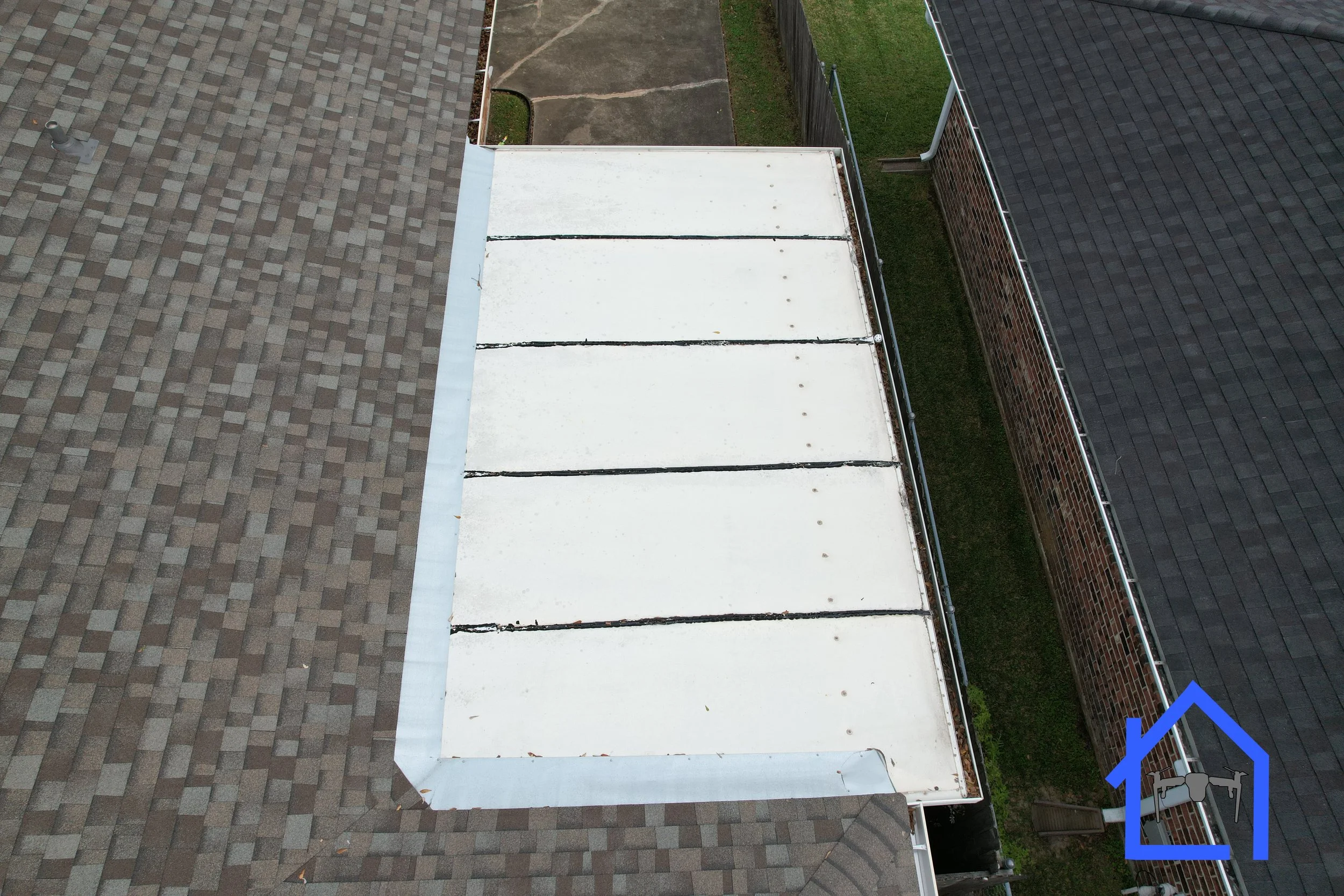 Aerial view of two house roofs with varying materials. The left roof is covered with dark shingles, and the right roof has a white flat section, possibly a garage or patio cover, adjacent to brown shingles.