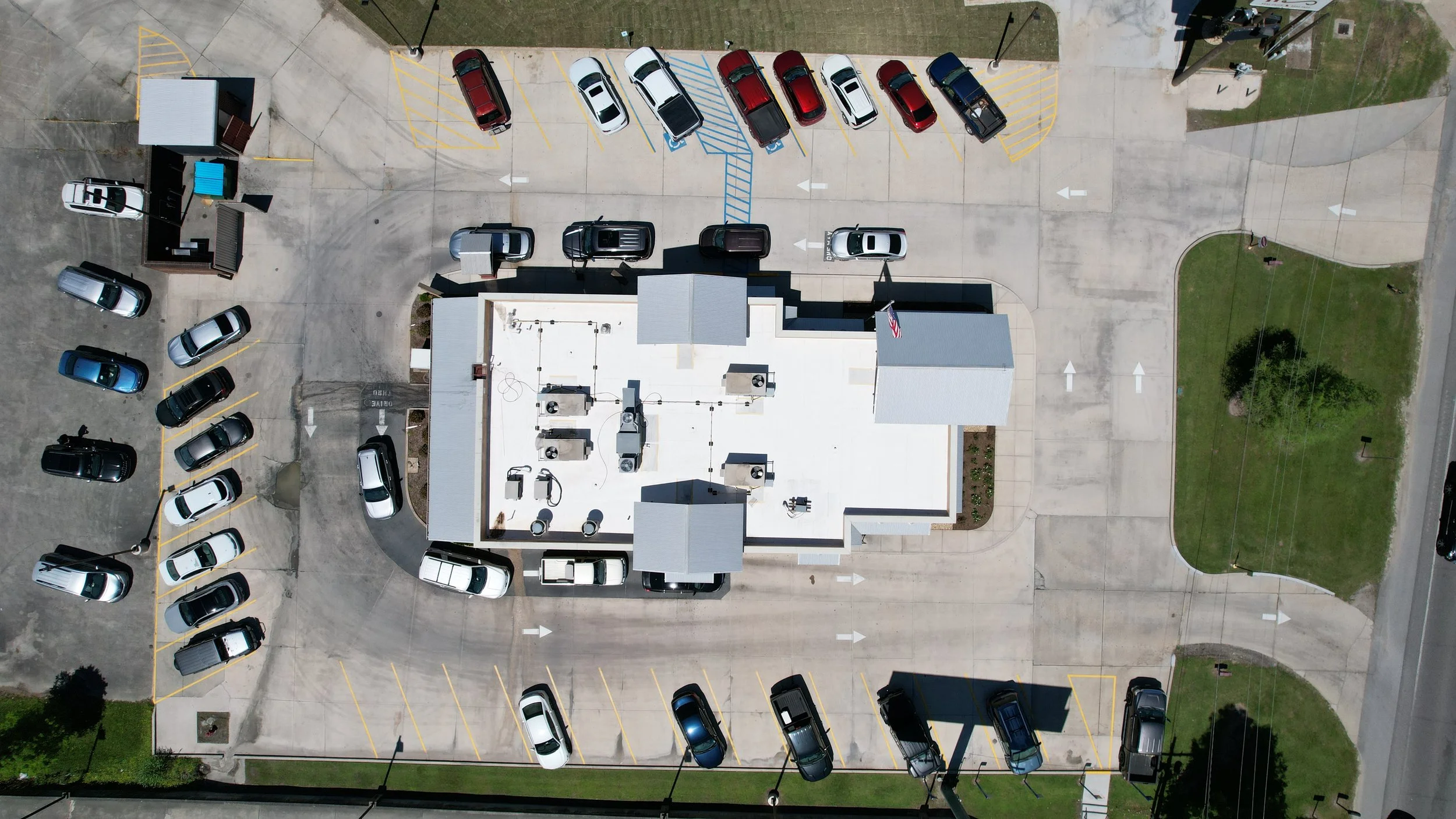 Aerial view of a parking lot with cars parked along the perimeter and in designated spaces, adjacent to a building with a roof and rooftop HVAC units. There are small green areas and sidewalks nearby.