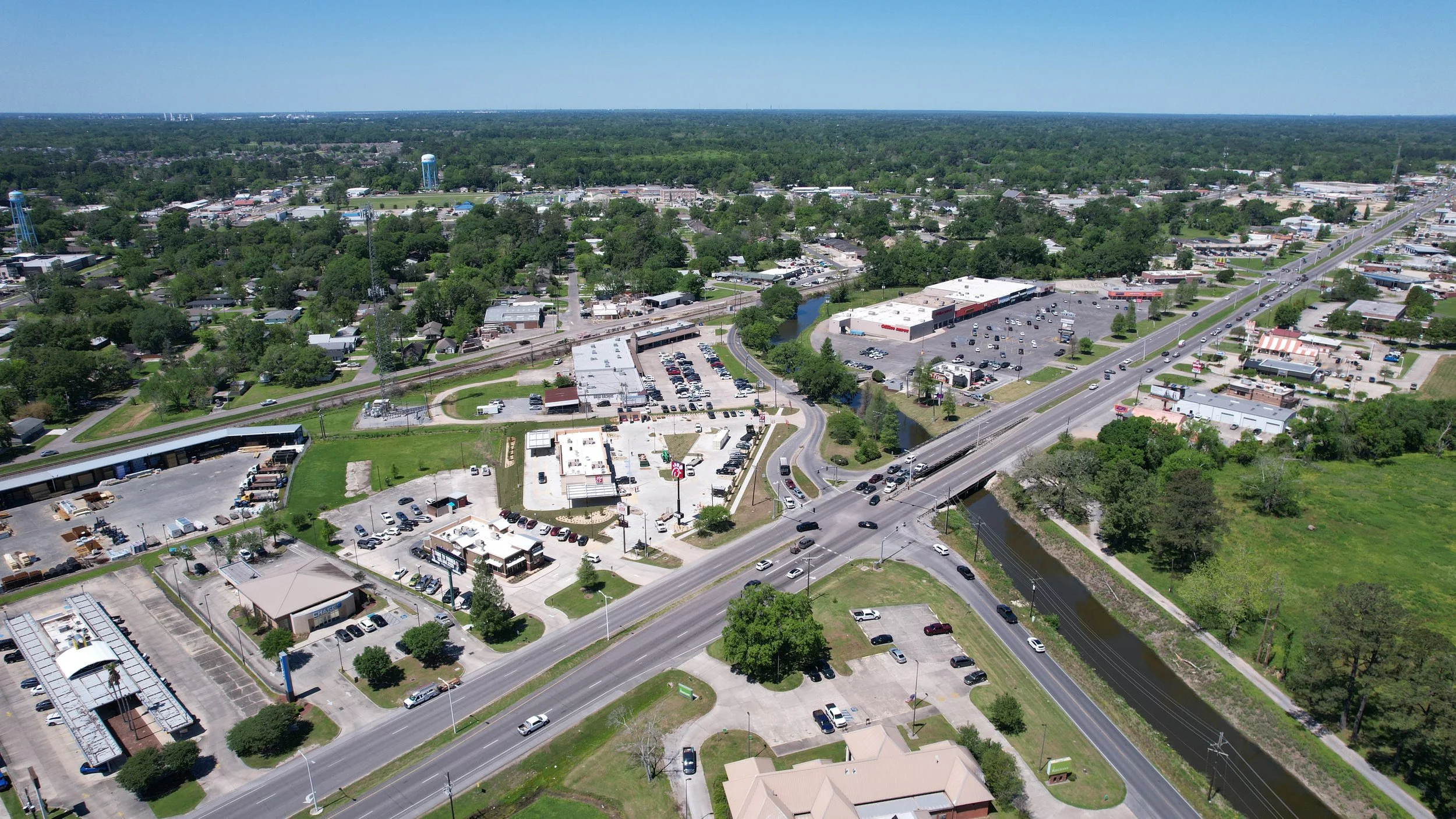 An aerial view of  Gonzales, Louisiana with commercial buildings along Blues Highway, Airlines Highway 61, a water tower, a canal crossing under the highway along a road and a residential neighborhood surrounded by trees under a clear blue sky.
