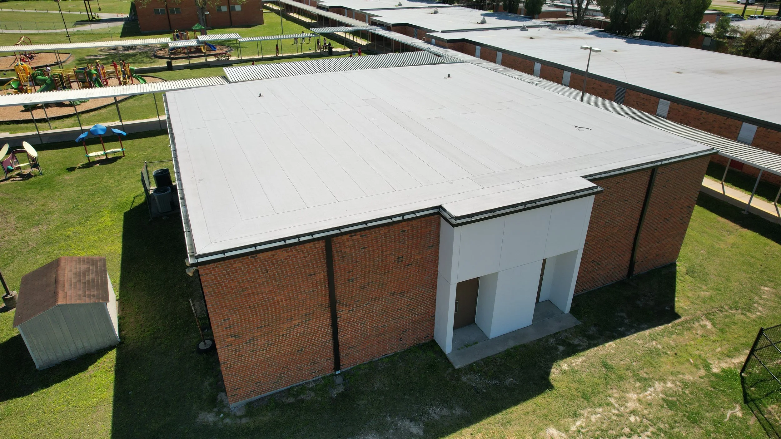 An aerial view of a school building with a new flat white roof installed by Supreme Roofing and Construction of Kenner, Louisiana and a playground with colorful equipment on a grassy area.