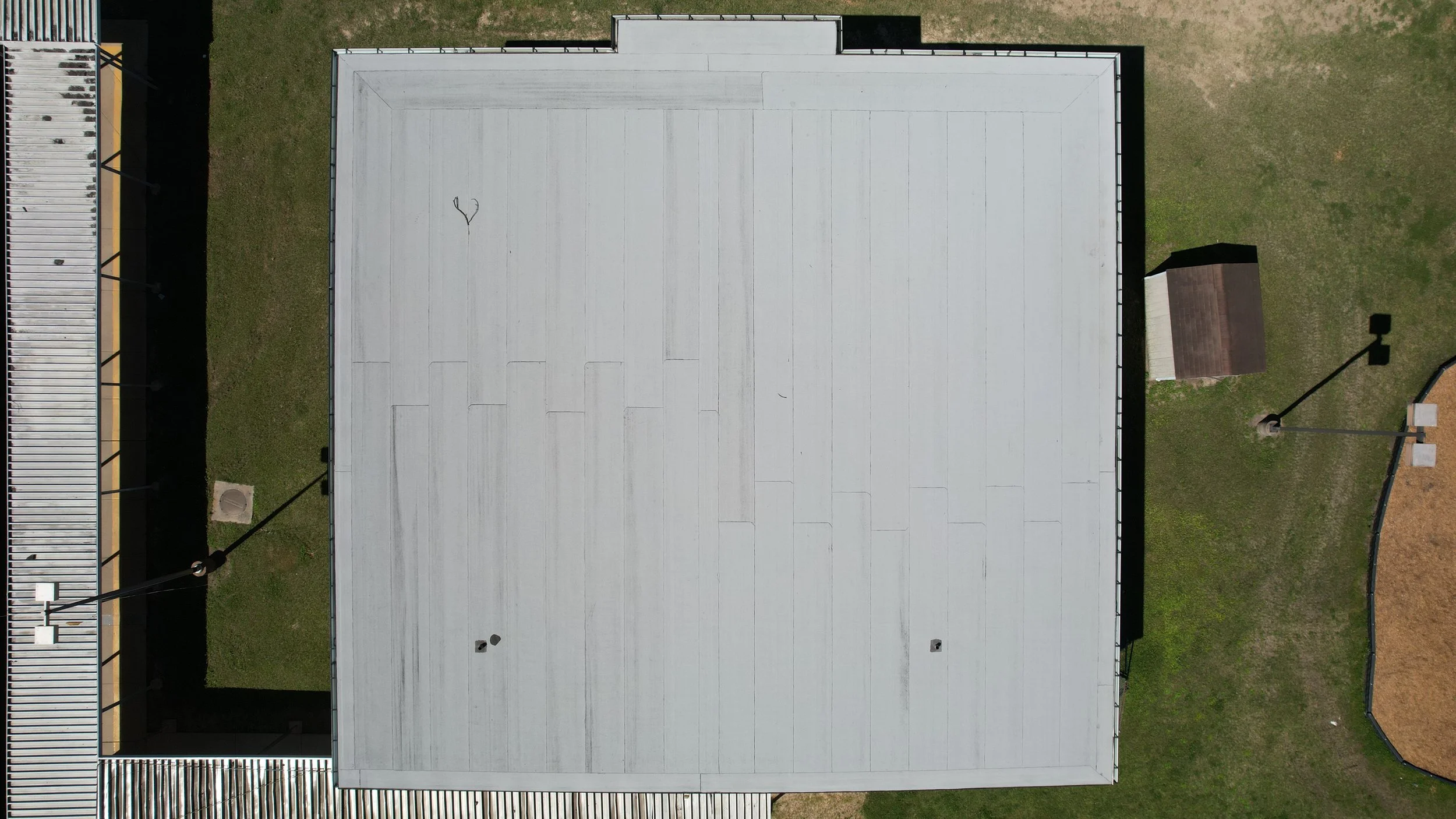 Aerial nadir view of a flat, white roof of a building with some black roof penetrations and two small vents, surrounded by grass, sidewalk, and outdoor structures.