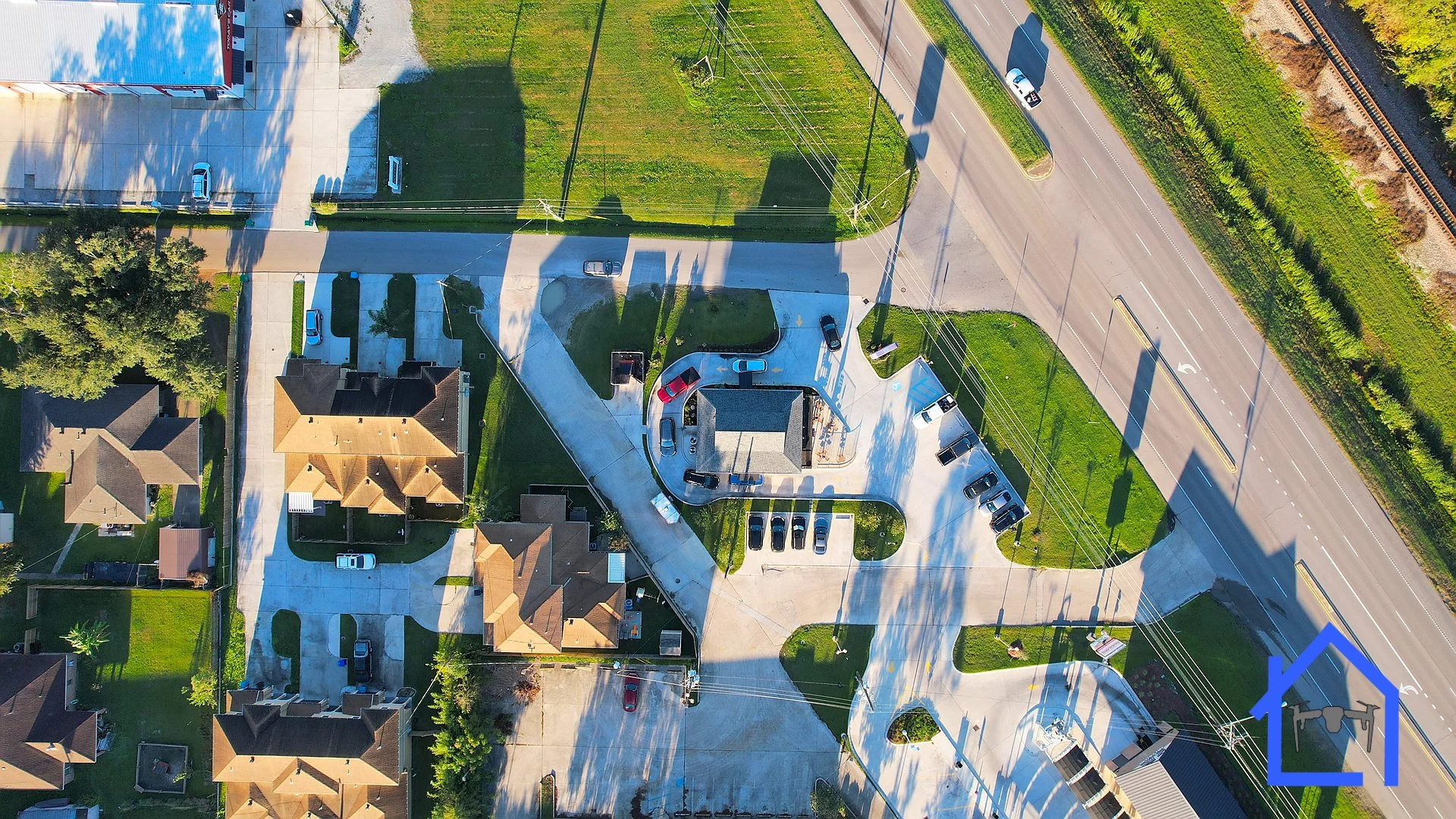 An aerial view of a residential neighborhood with houses, driveways, parked cars, and a small playground. A major road runs along the right edge of the image.