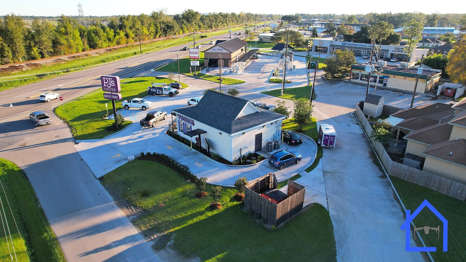 Aerial view of a coffee shop with a drive-thru, parking lot, and surrounding roads in a suburban area, with houses, trees, and commercial buildings.