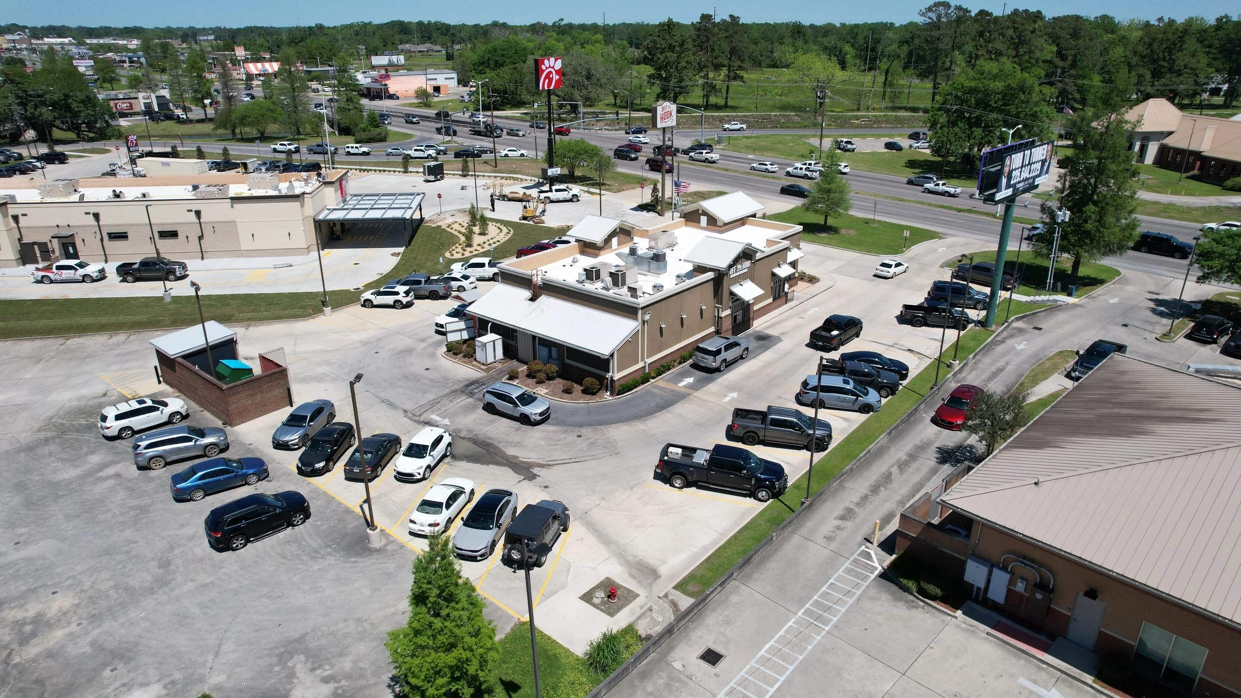 Aerial view of a parking lot and fast food Chick-Fil-A restaurant under construction with a drive-thru, surrounded by trees, roads, and commercial buildings.