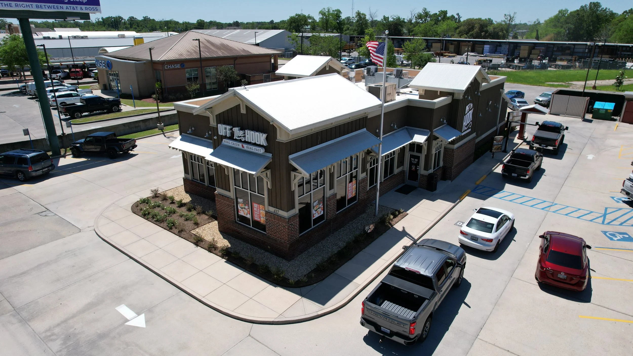 Aerial view of a busy highway with many place to shop, eat and play with a restaurant called 'Off The Hook', cars parked in parking lot, Chase bank building in background, American flag flying, and surrounding greenery.
