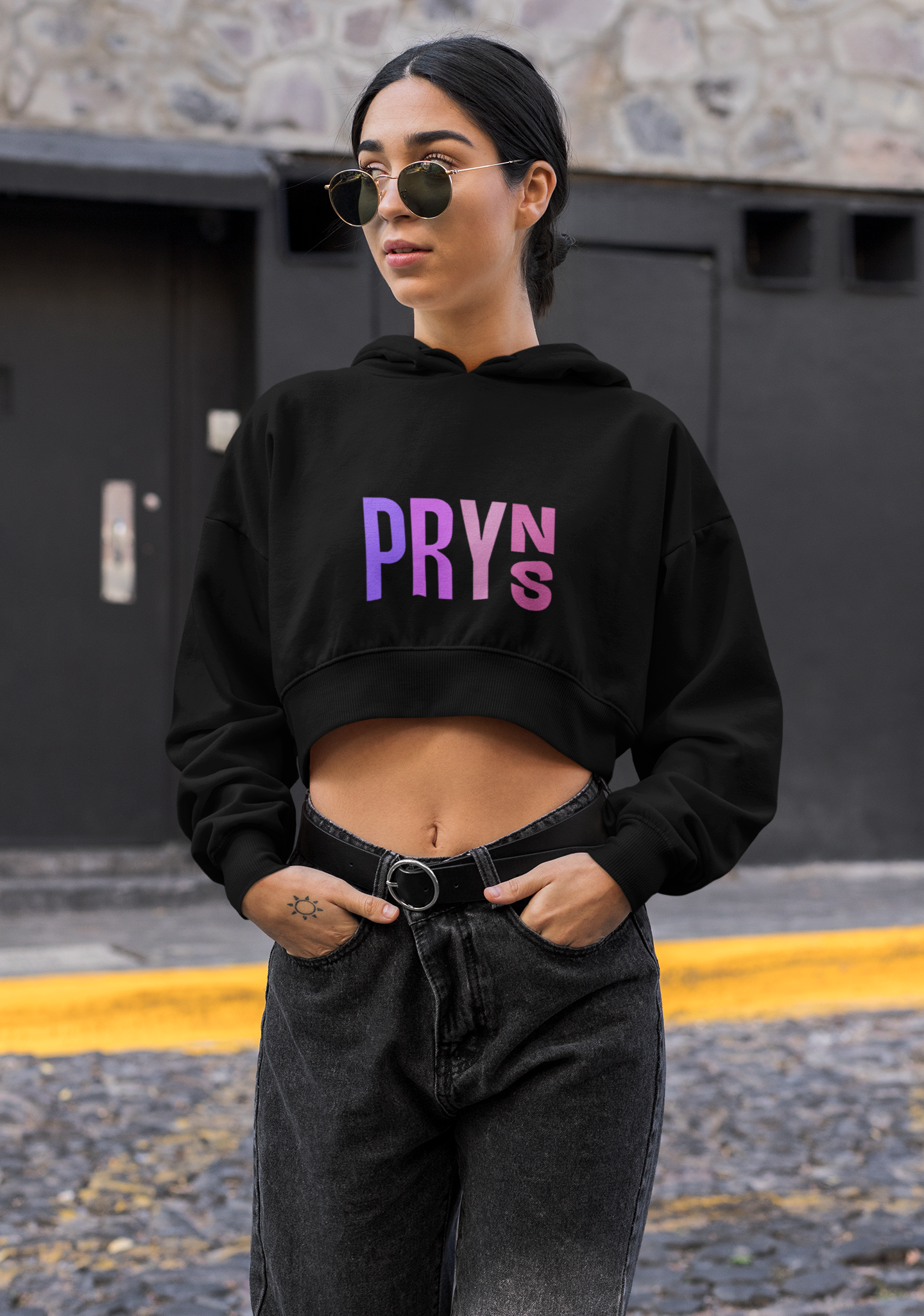 Fashionably dressed woman standing outdoors in front of a dark wall, wearing round sunglasses, a black cropped hoodie with purple and pink text, black jeans, and a black belt.