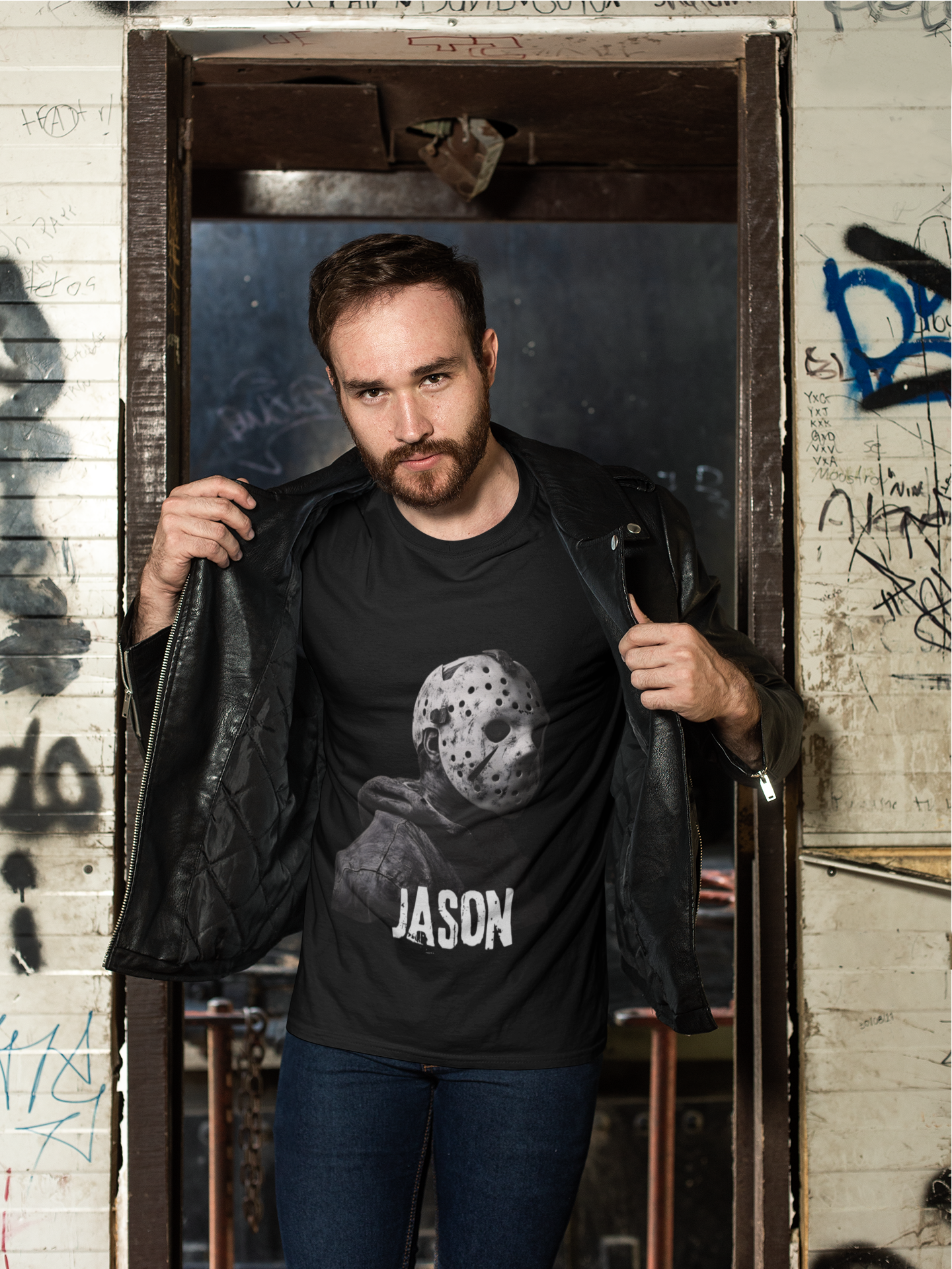 A man with a beard and short hair leans in a doorway with graffiti-covered walls behind him. He is wearing a black leather jacket and a black T-shirt featuring a Fox Michael Myers mask and the name 'Jason' underneath. He is holding his jacket open with both hands.