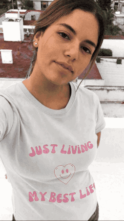 Young woman with brown hair in a ponytail taking a selfie outdoors, wearing a white T-shirt with pink text that says 'Just Living My Best Life' and a smiley face.