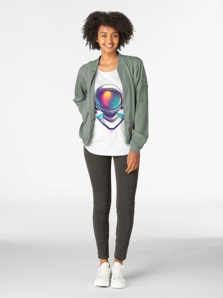 Young woman smiling, wearing a green bomber jacket, graphic astronaut t-shirt, gray jeans, and white sneakers, standing against a white background.