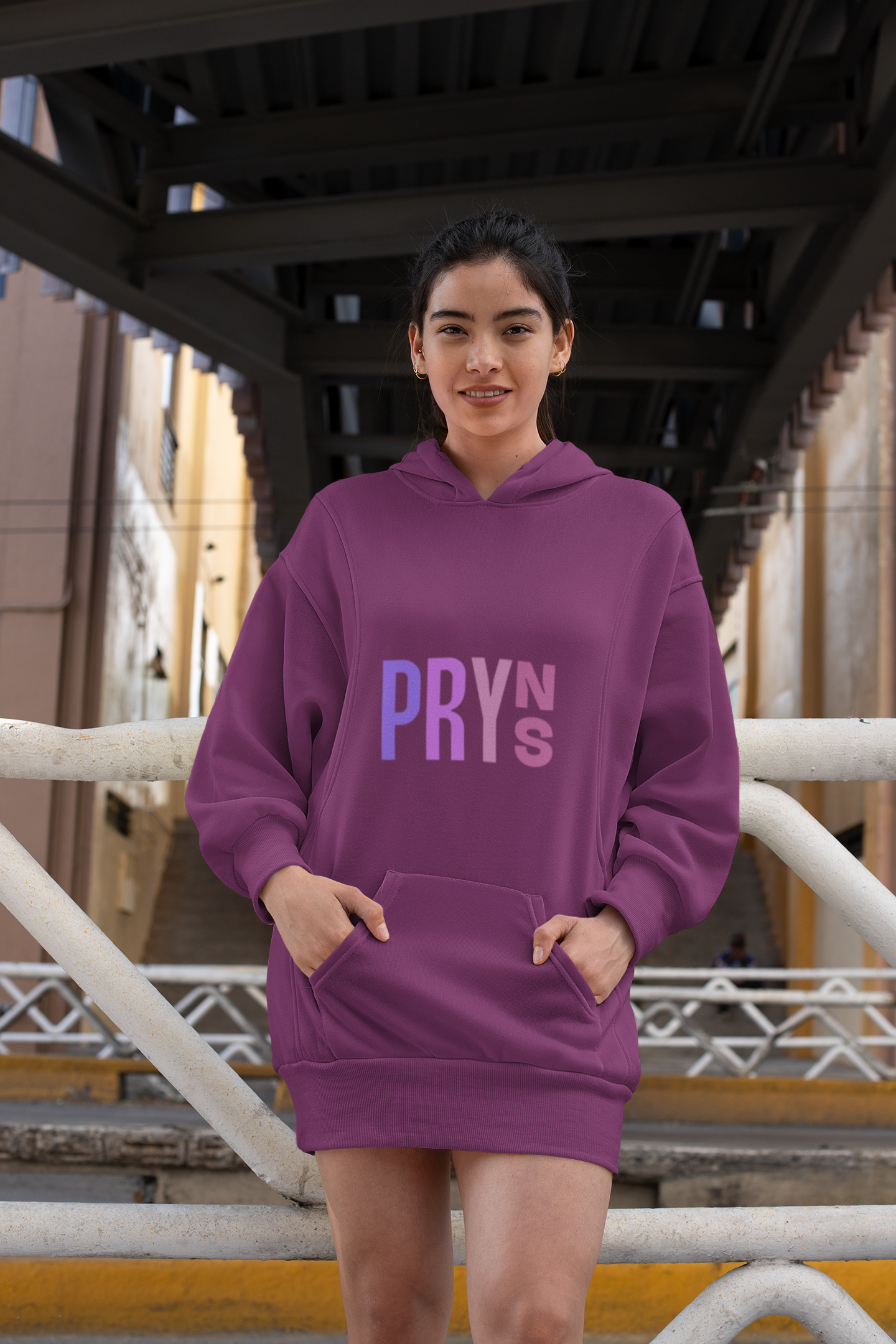 Young woman in a purple hoodie with text design, standing outdoors under a dark metal staircase, with a yellow building in the background.