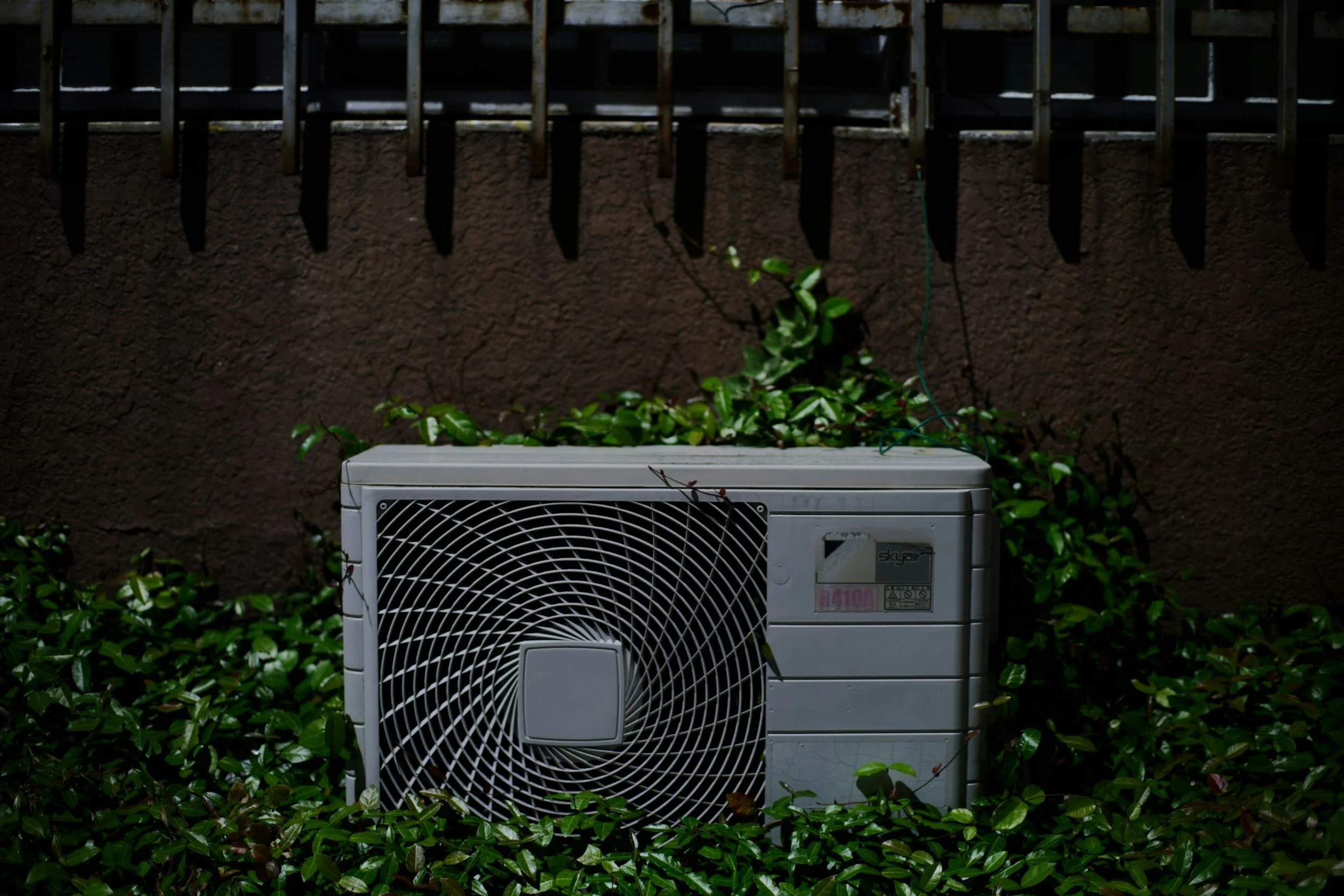 Outdoor scene with an air conditioning unit placed among green bushes, with a brown wall and a metal balcony above.
