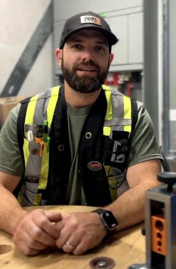 A man with a beard wearing a black cap, gray shirt, and a yellow safety vest with tools. Sitting at a wooden table in a workshop or industrial setting.