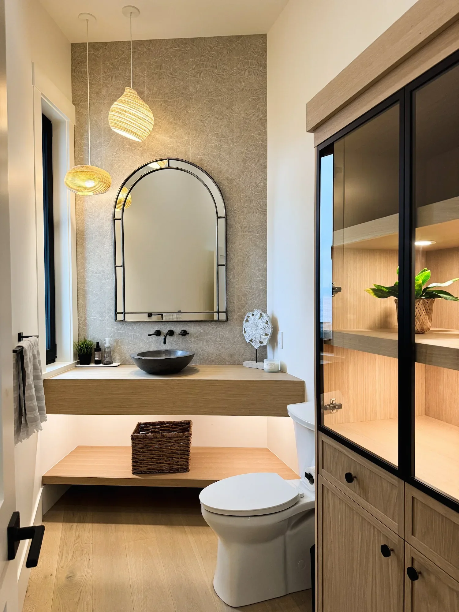 Modern bathroom with two hanging pendant lights, a round mirror, a vessel sink, plant decor, a wooden countertop, and a toilet, with a wooden cabinet and a basket underneath.