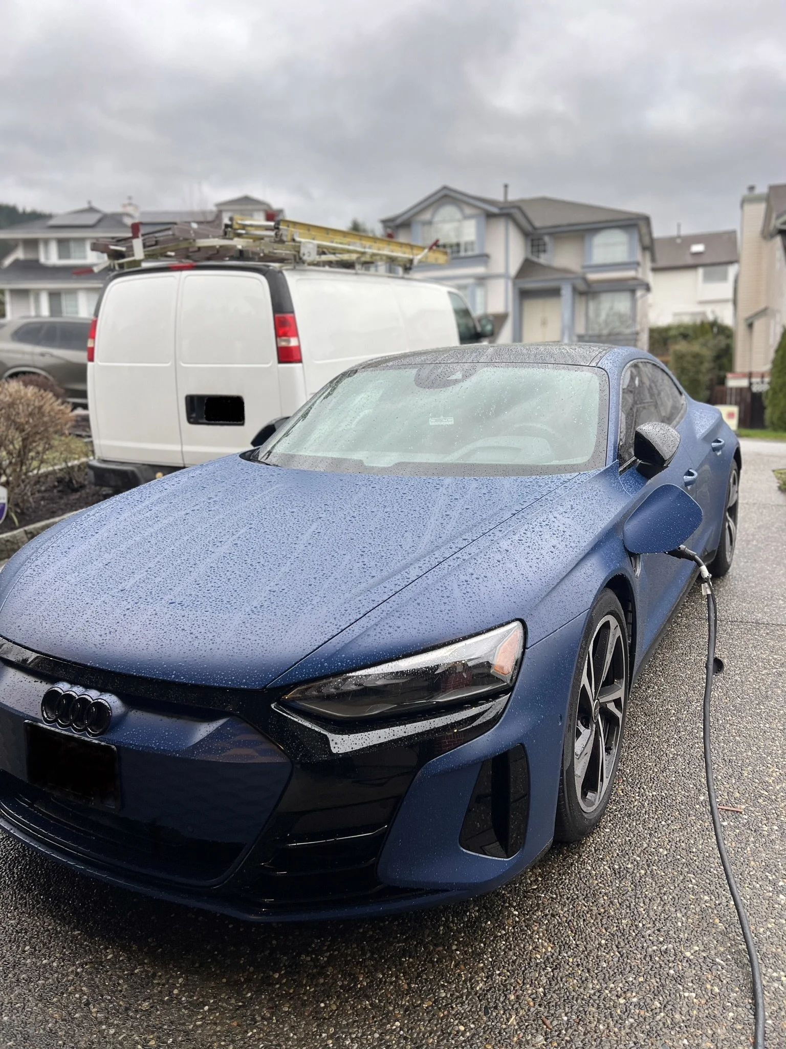 A EV car parked outdoors, connected to an electric charger, with rain drops on its surface, and residential houses in the background on a cloudy day.
