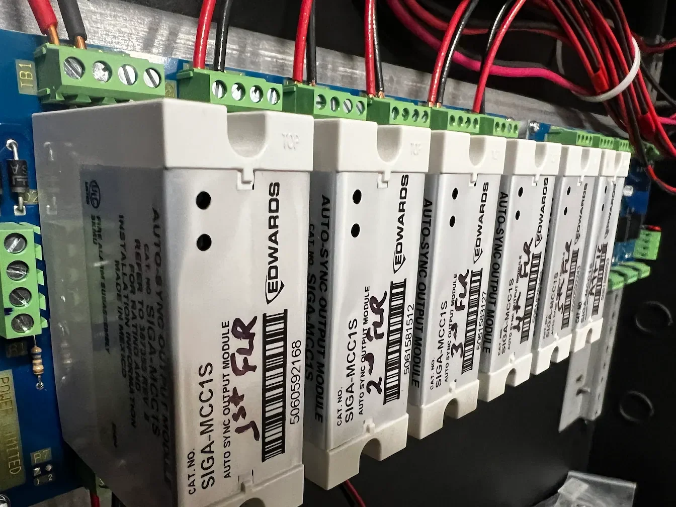 Multiple white signal transponder modules with black text and barcodes, mounted on an electrical panel, with green terminal blocks connected to red and black wires.