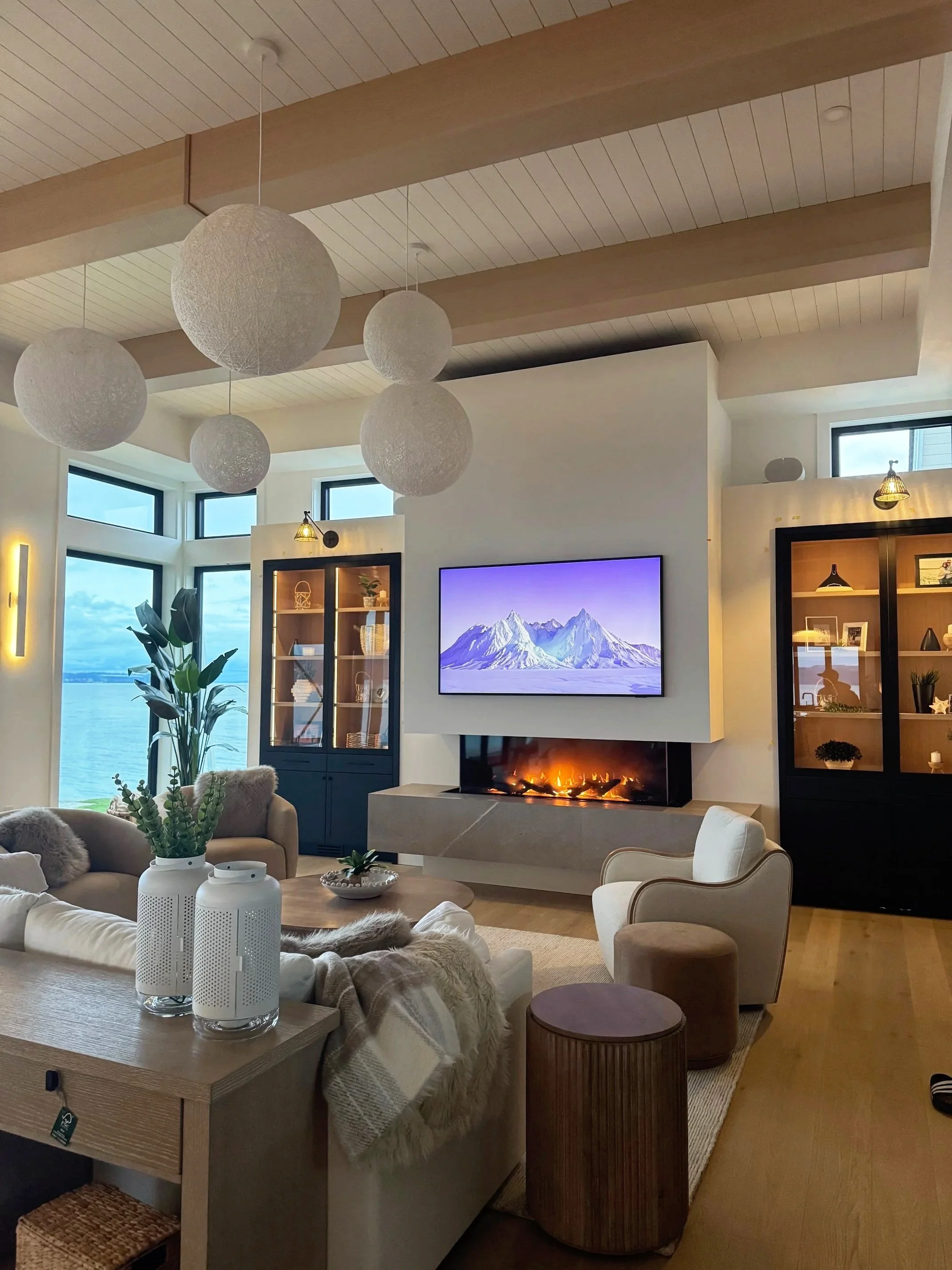 A modern living room with floor-to-ceiling windows overlooking water. It features a mounted flat-screen TV above a fireplace, neutral-colored furniture, wooden ceiling beams with hanging round paper lanterns, and decorative shelves.