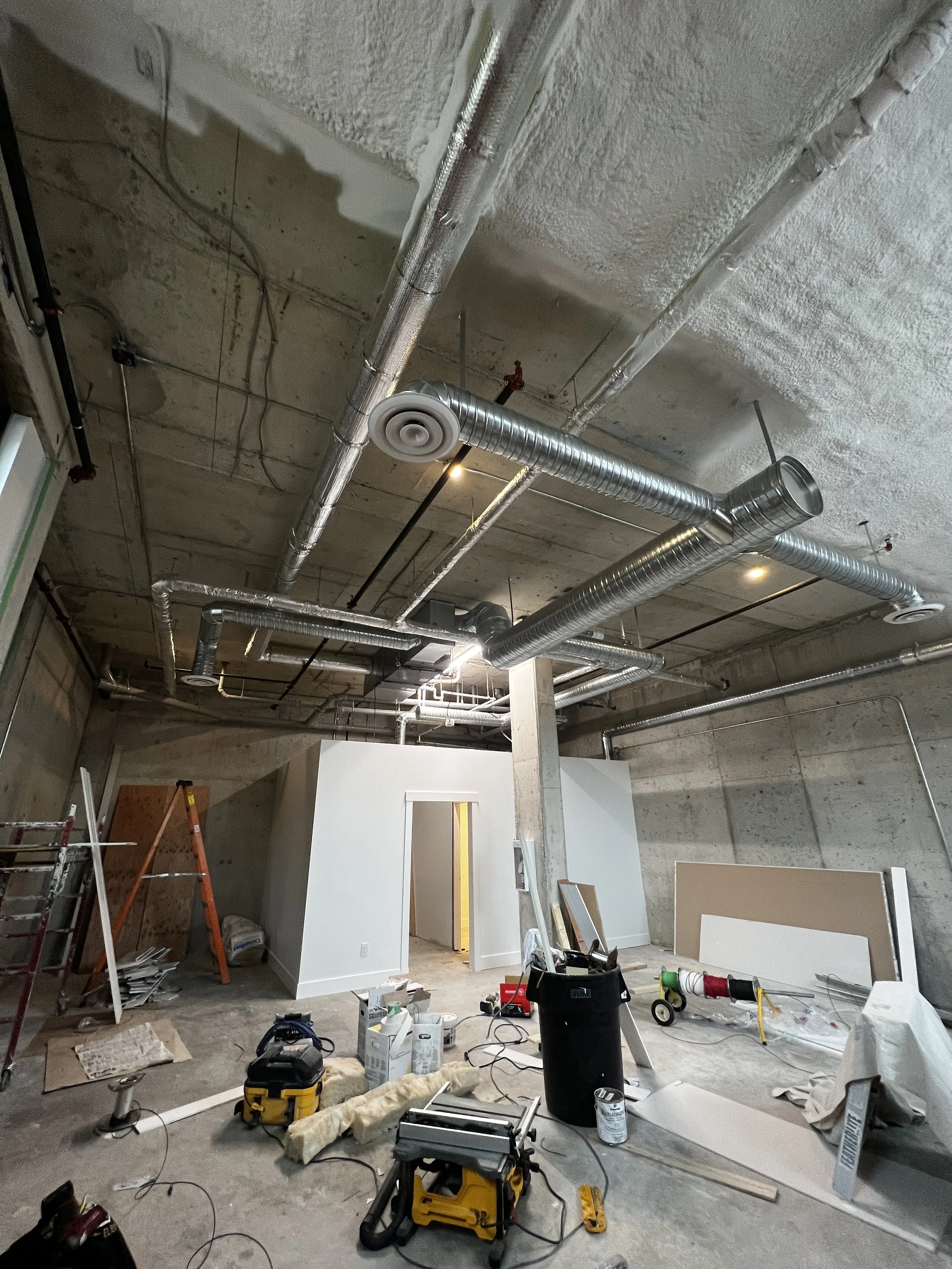 Construction site in progress inside an unfinished building, with exposed ductwork on the ceiling, tools, and materials scattered on the floor.