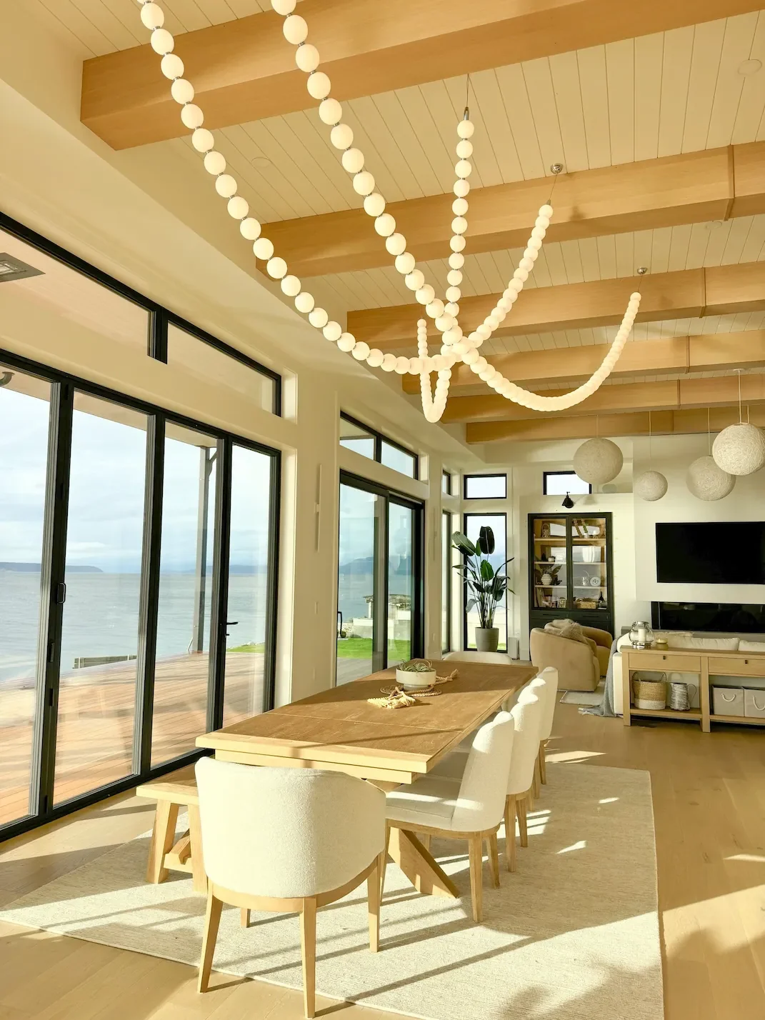 Bright living and dining area with large windows overlooking water, wooden ceiling beams, modern white chandelier, dining table with chairs, sofa, TV, and houseplants.