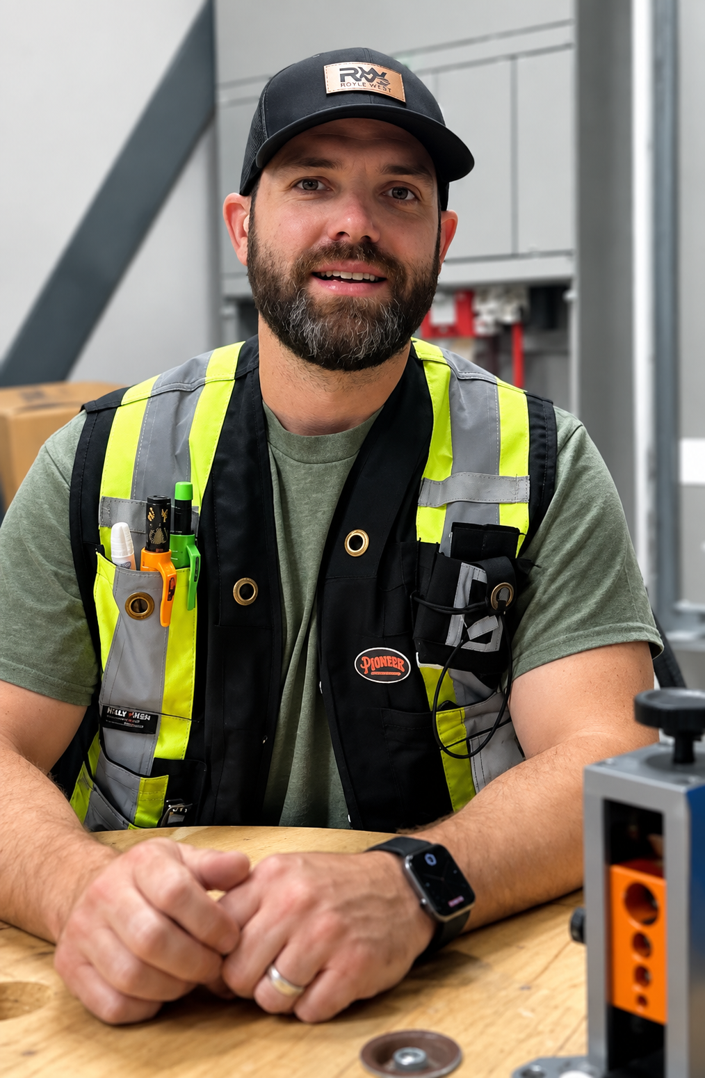 A smiling man with a beard, wearing a black cap, a gray shirt, and a neon yellow and black safety vest, sitting at a workbench with tools in his vest pocket.