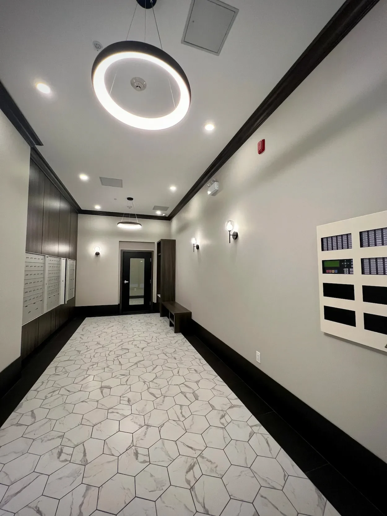 Modern building lobby with white hexagonal marble flooring, black trim, wall-mounted lights, mailboxes on the left, and a door at the end of the hall.