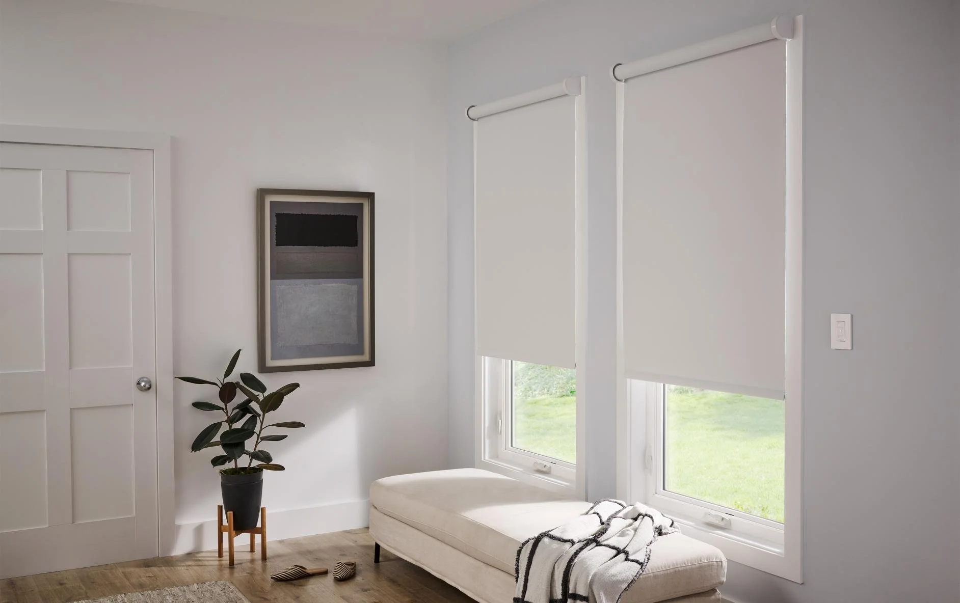 A minimalist interior with a white bench under two large windows with white roller shades, a potted plant on a small wooden stand, a framed abstract art piece on the wall, and a pair of striped slippers on the wooden floor.