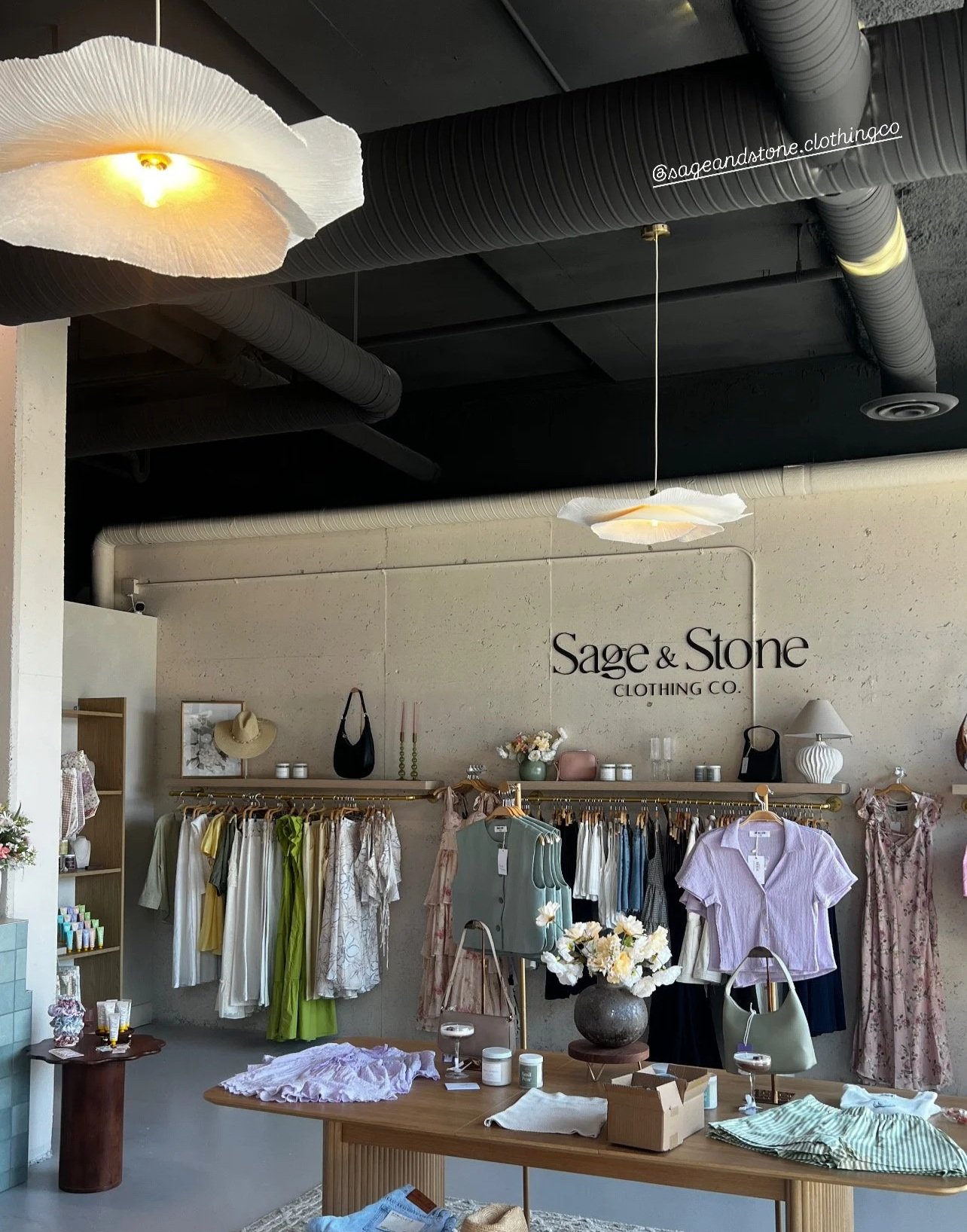 Inside a clothing store named Sage & Stone, displaying women's clothing, accessories, and decorative items on racks and shelves, with stylish lighting and a simple, elegant decor.