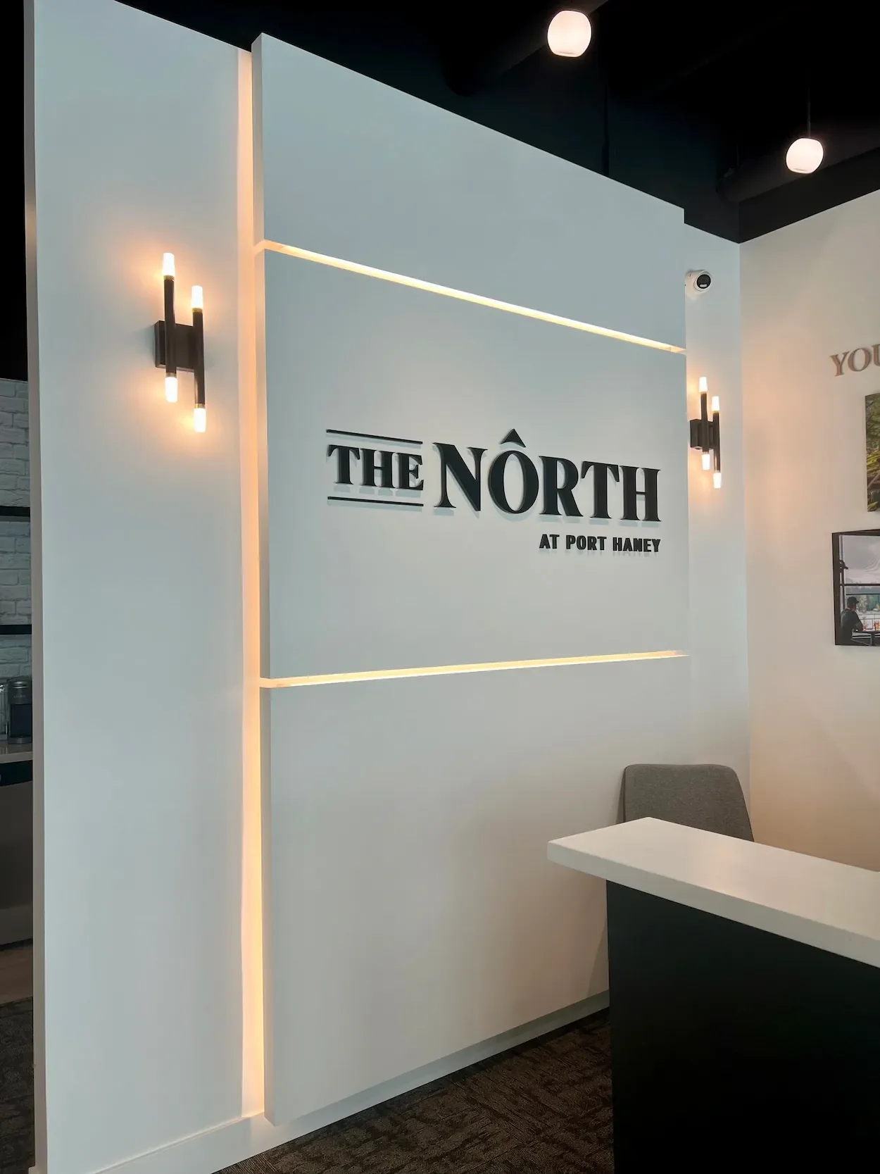 Interior of a modern building with a white wall displaying the sign 'The North at Port Hueny'. The wall features black letter signage and illuminated wall sconces, with a gray chair and a white table nearby.