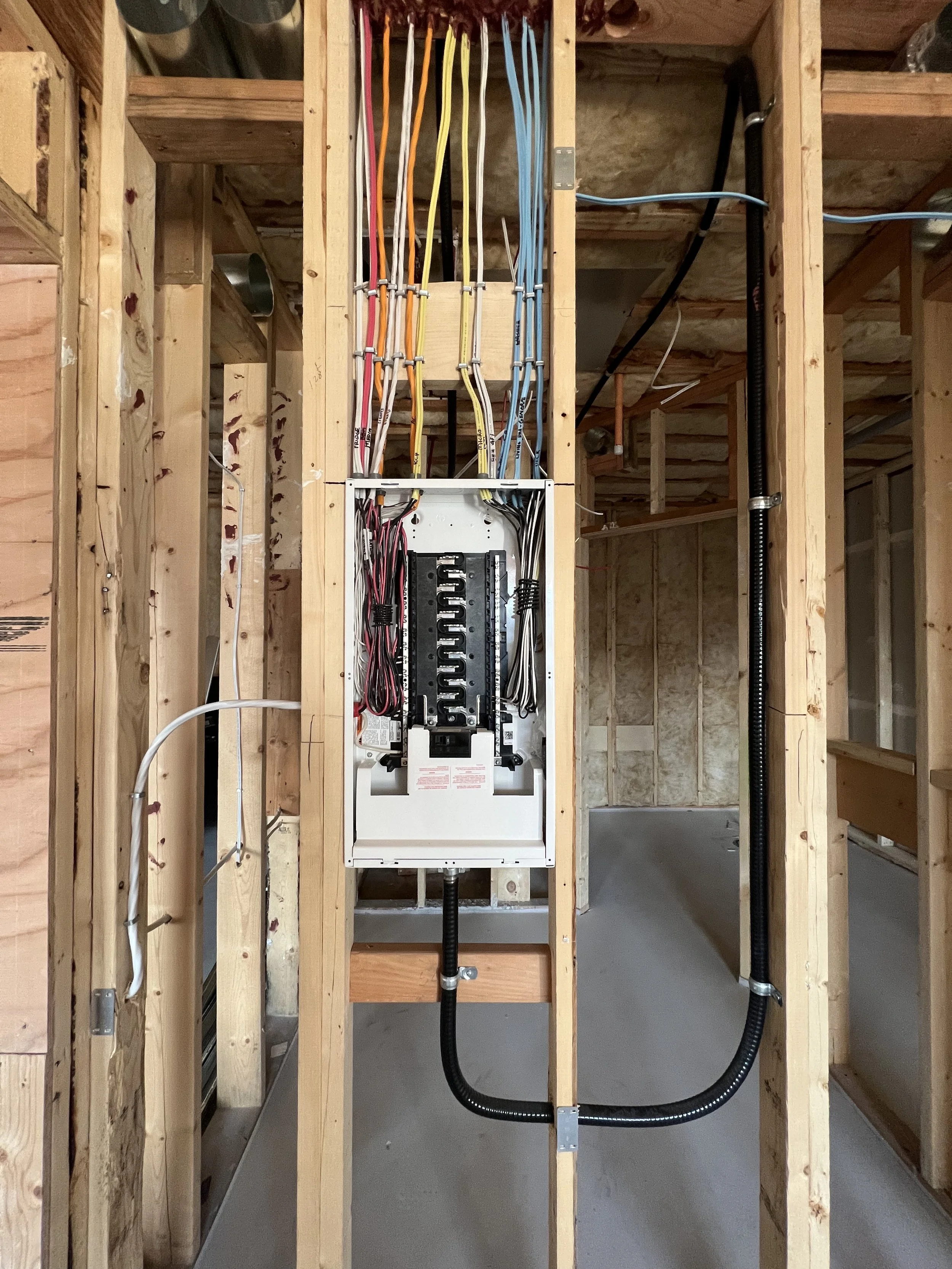 Electrical panel with multiple colored wires installed in a wooden-framed room under construction.