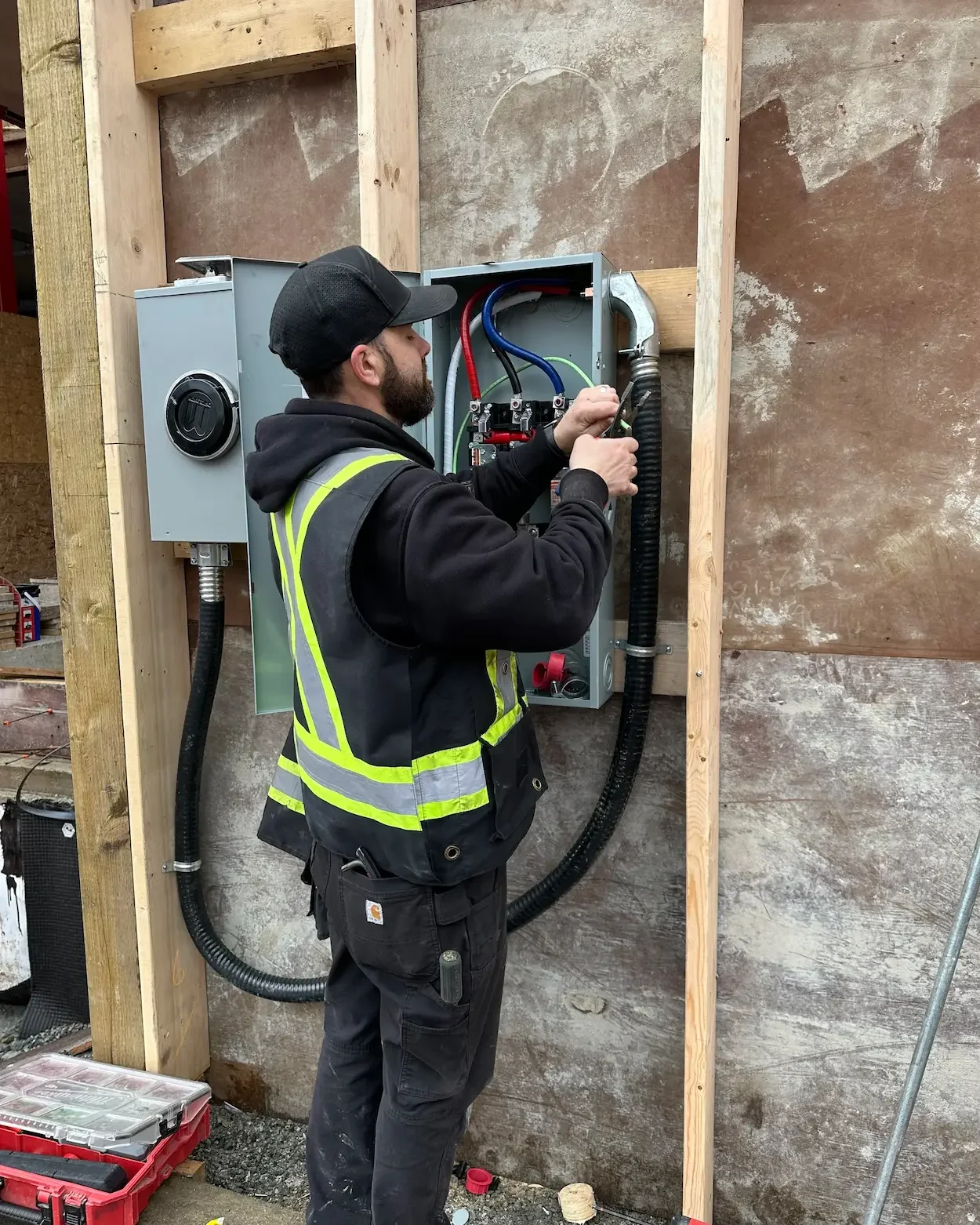 A man working on an electrical panel outdoors, wearing a black cap, black hoodie, and a yellow safety vest, with tools and equipment nearby.